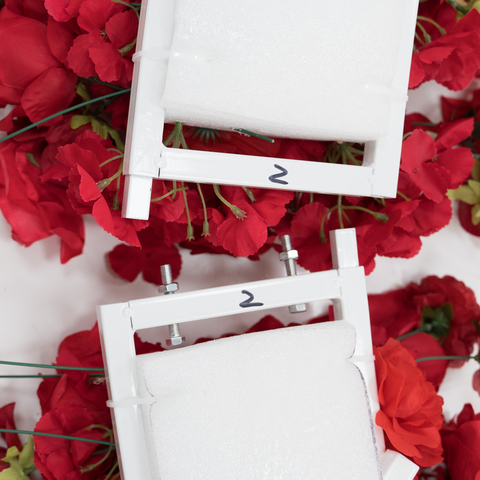 Close - up of two white metal frames labeled “2”, each with foam padding. They’re surrounded by red artificial flowers, likely part of a decorative floral structure. The frames show assembly components like bolts, suggesting they’re for building a larger decorative piece. 