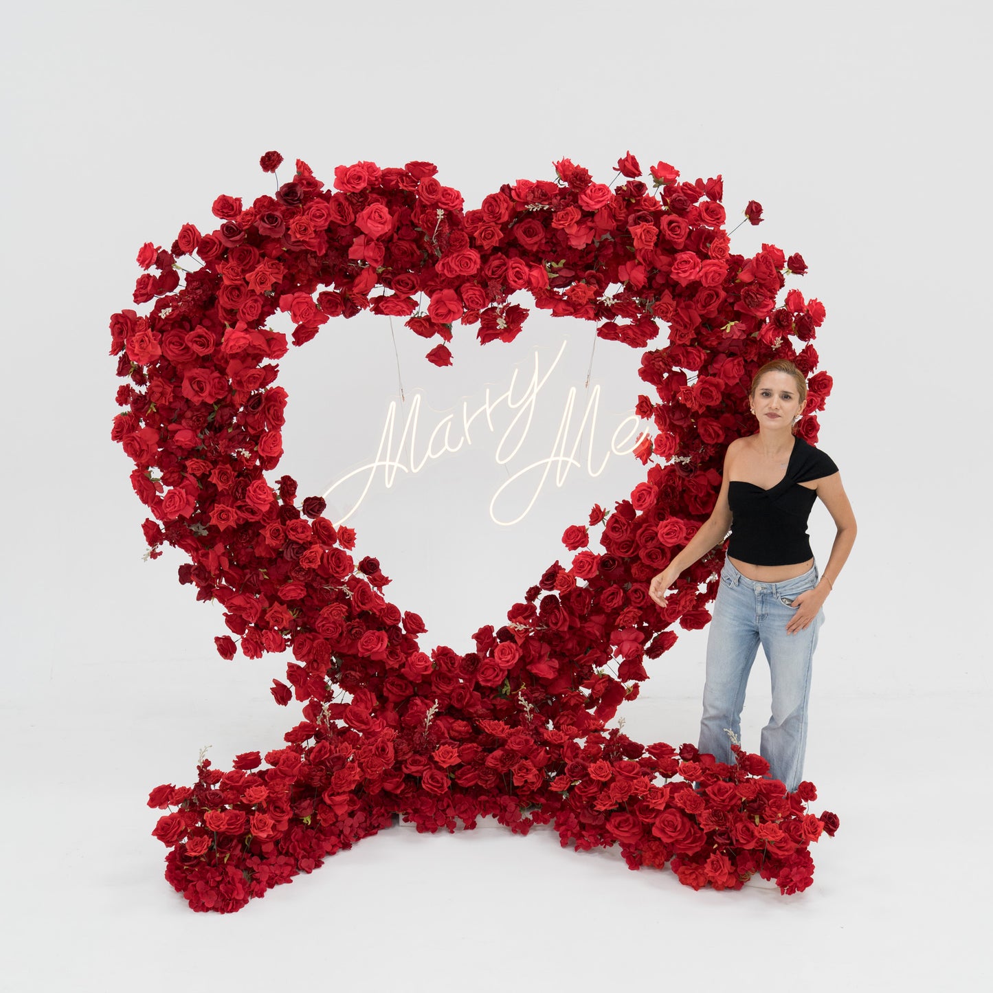 A woman stands beside a huge heart - shaped red rose arrangement. In the center, a glowing “Marry Me” sign adds romance. The vibrant blooms and her casual black top - and - jeans look create a sweet, memorable proposal - style scene. 