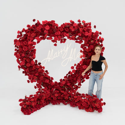 A woman stands beside a huge heart - shaped red rose arrangement. In the center, a glowing “Marry Me” sign adds romance. The vibrant blooms and her casual black top - and - jeans look create a sweet, memorable proposal - style scene. 
