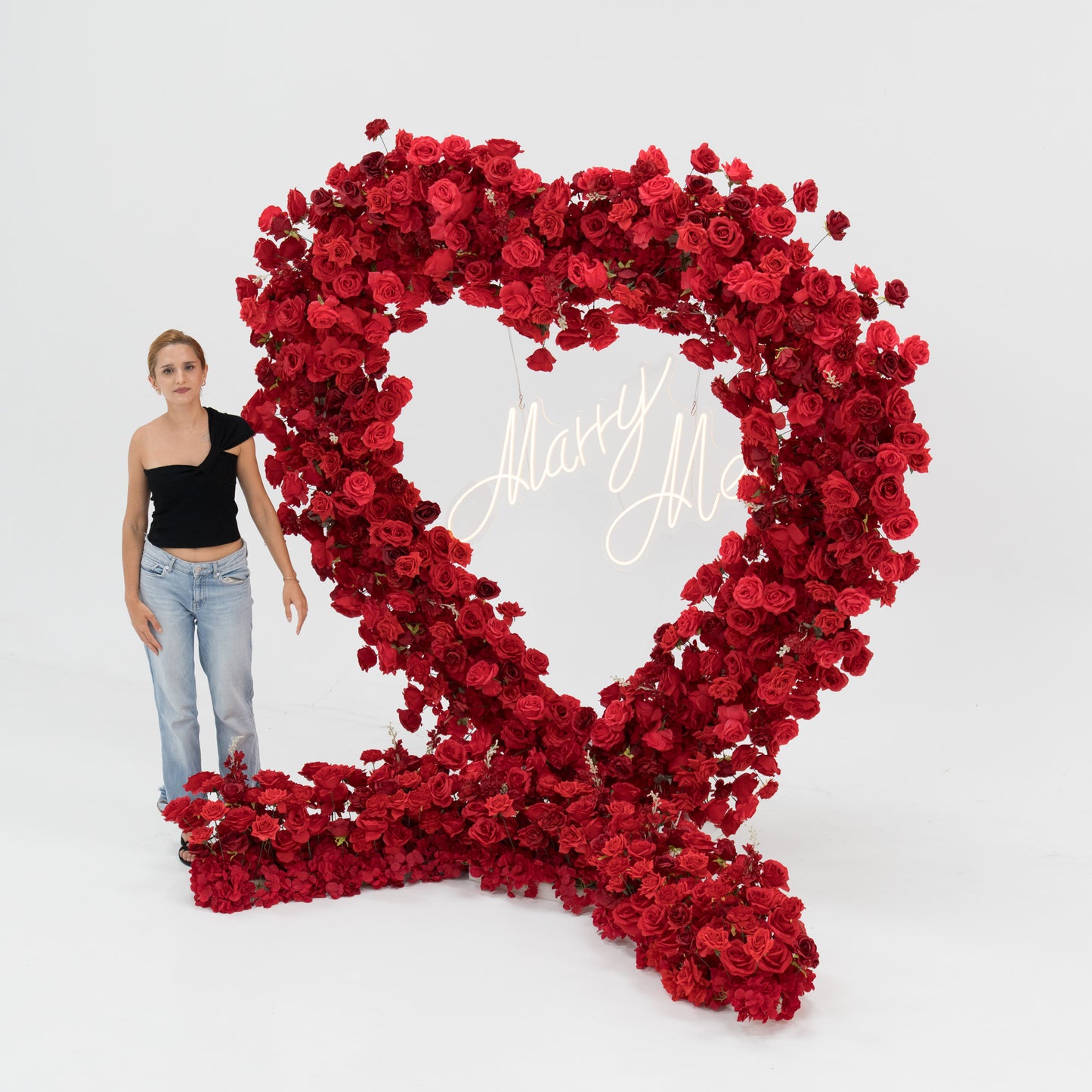 A woman in a black one - shoulder top and blue jeans stands beside a massive heart - shaped red rose installation. At its center, a glowing “Marry Me” sign adds a romantic touch. The vibrant, lush blooms create a striking, proposal - worthy scene. 