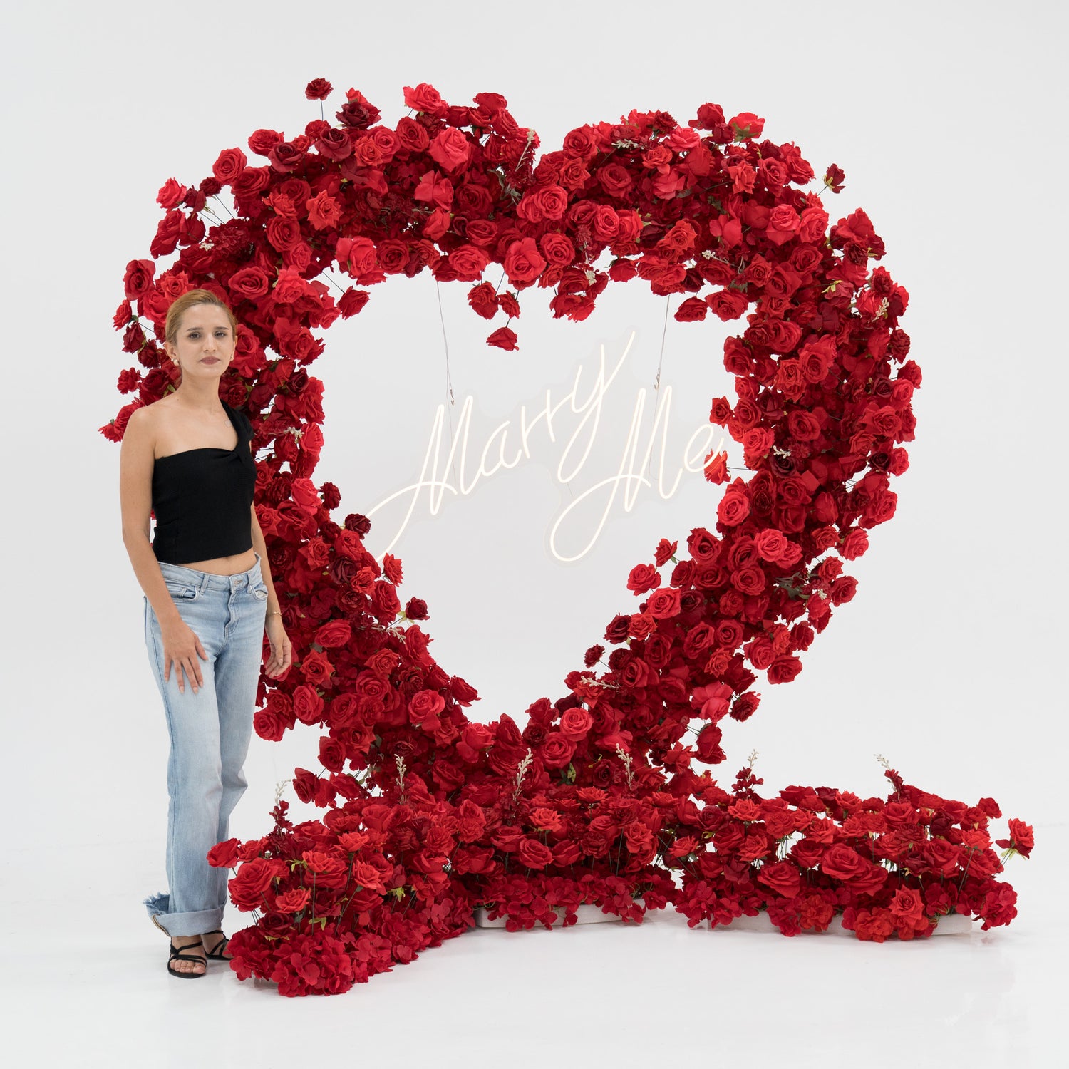A woman in a black one - shoulder top and blue jeans stands beside a large heart - shaped red rose installation. At its center, a glowing “Marry Me” sign adds a romantic touch. The lush, vibrant blooms create a striking, proposal - worthy scene. 