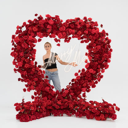 A woman stands within a large heart - shaped red rose installation. At the center, a glowing “Marry Me” sign adds romance. She wears a black one - shoulder top and blue jeans, with arms outstretched, creating a sweet, proposal - like scene filled with vibrant blooms. 