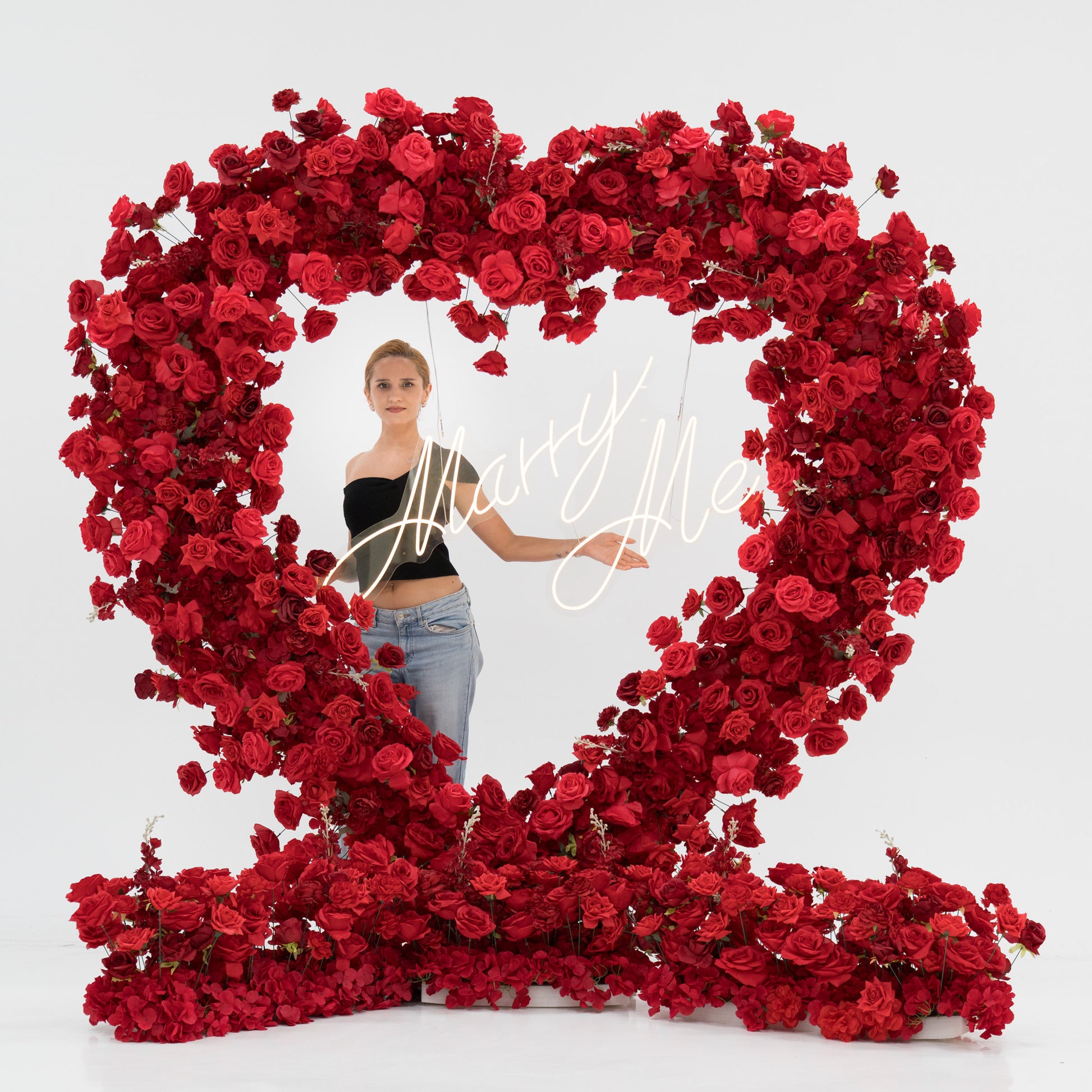 A woman stands inside a huge heart - shaped red rose installation. A glowing “Marry Me” sign hangs in the center. She wears a black one - shoulder top and blue jeans, with arms outstretched, creating a romantic and memorable proposal - style scene. 