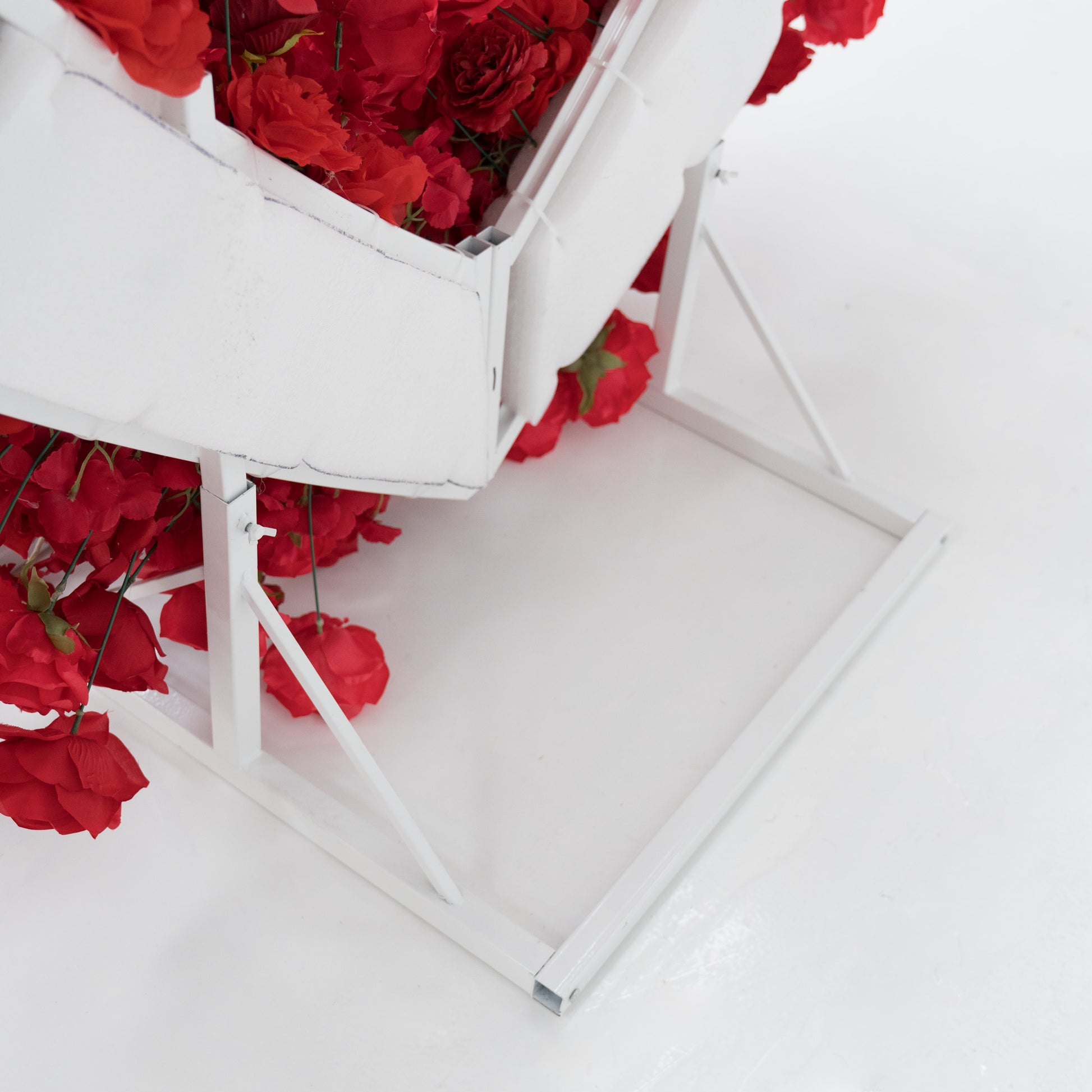 This is a close - up of a white metal frame structure, likely part of a decorative flower display. Red artificial roses spill from it, contrasting with the white frame and background. Simple yet elegant, it highlights the support system for floral decorations, blending functionality with visual appeal. 