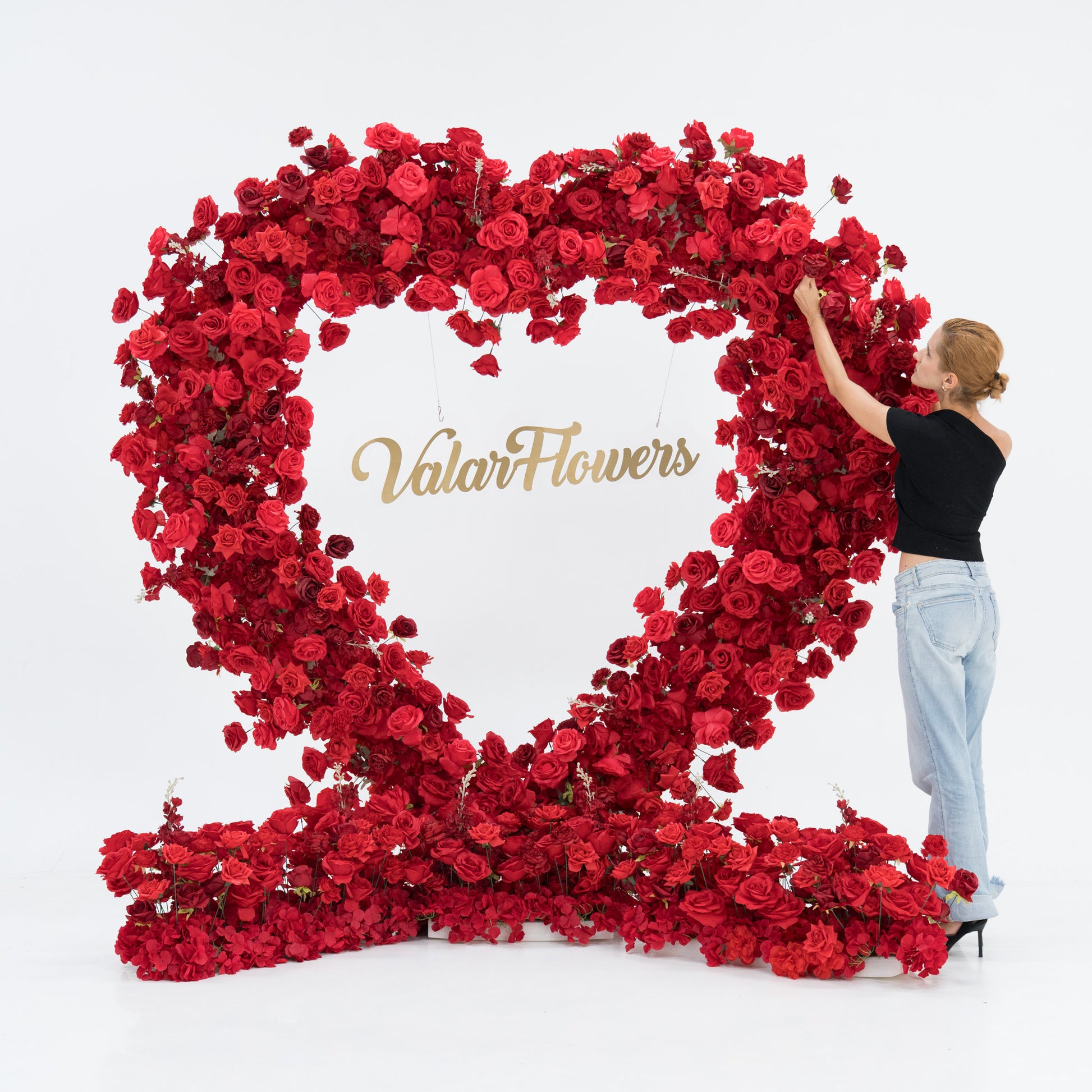A large heart - shaped red rose installation with “ValarFlowers” in gold at the center. A woman in a black top and blue jeans adjusts the blooms, adding a human touch to the lush, vibrant floral display, creating a romantic and artistic scene. 