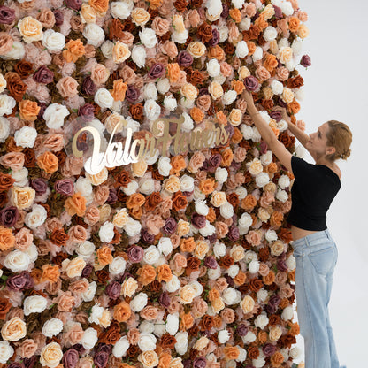 A woman in a black top and blue jeans adjusts a large floral backdrop. The backdrop features beige, white, and purple artificial flowers, with “ValarFlowers” in gold script. She adds a human touch to the elegant, textured display. 