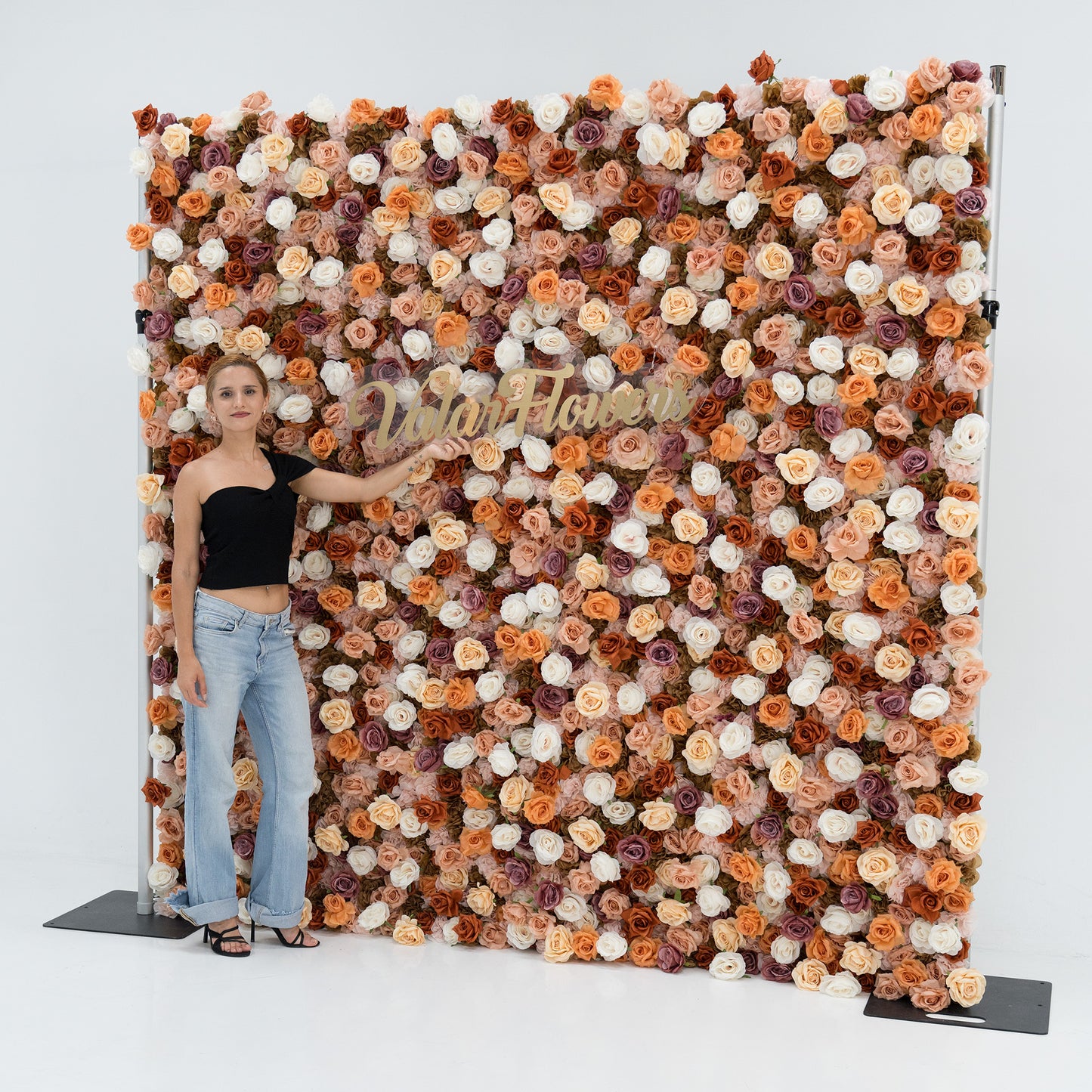 A woman in a black one - shoulder top and blue jeans stands beside a large floral backdrop. The backdrop features beige, white, and brown artificial flowers, with “ValarFlowers” in gold script. She poses, adding a human element to the elegant, textured display. 