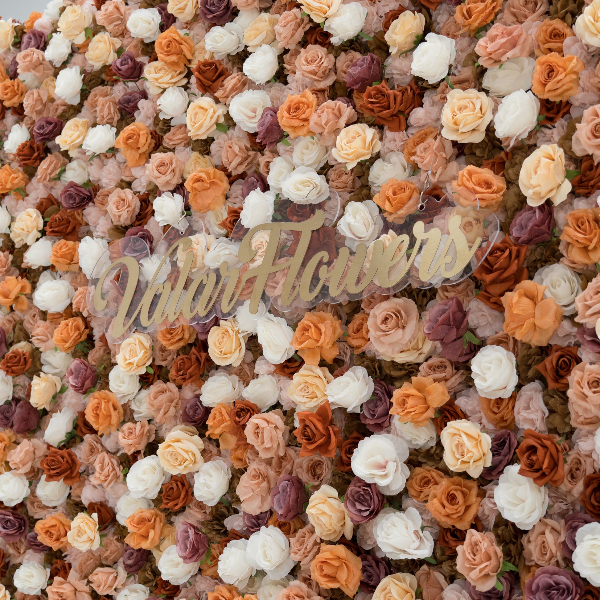 A close - up of a gold “ValarFlowers” sign against a lush artificial flower wall. The wall features beige, white, peach, and purple roses, creating a textured, elegant backdrop. The sign adds a branding element to the decorative floral display. 
