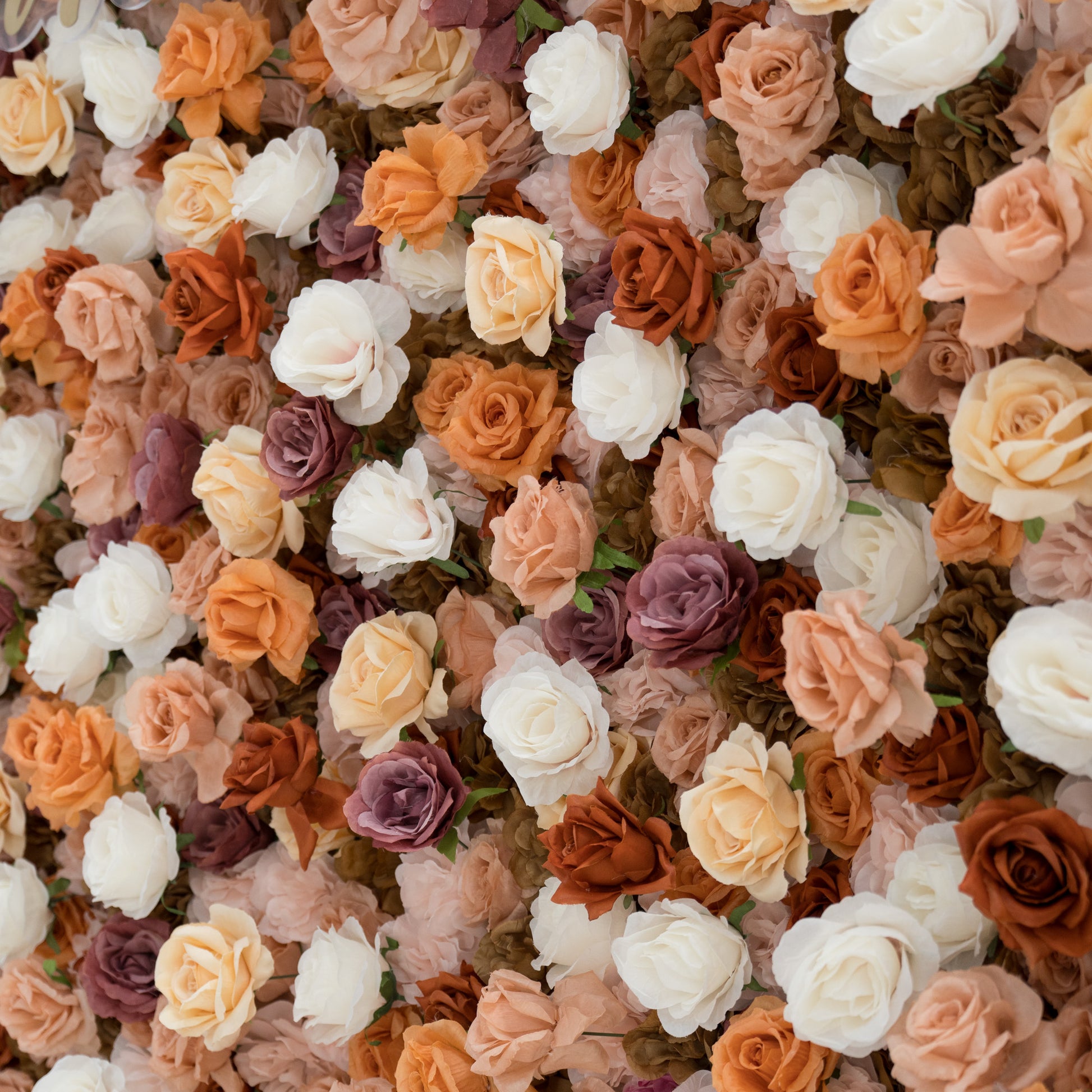A close - up of a gold “ValarFlowers” sign against a lush artificial flower wall. The wall features beige, white, peach, and purple roses, creating a textured, elegant backdrop. The sign adds a branding element to the decorative floral display. 