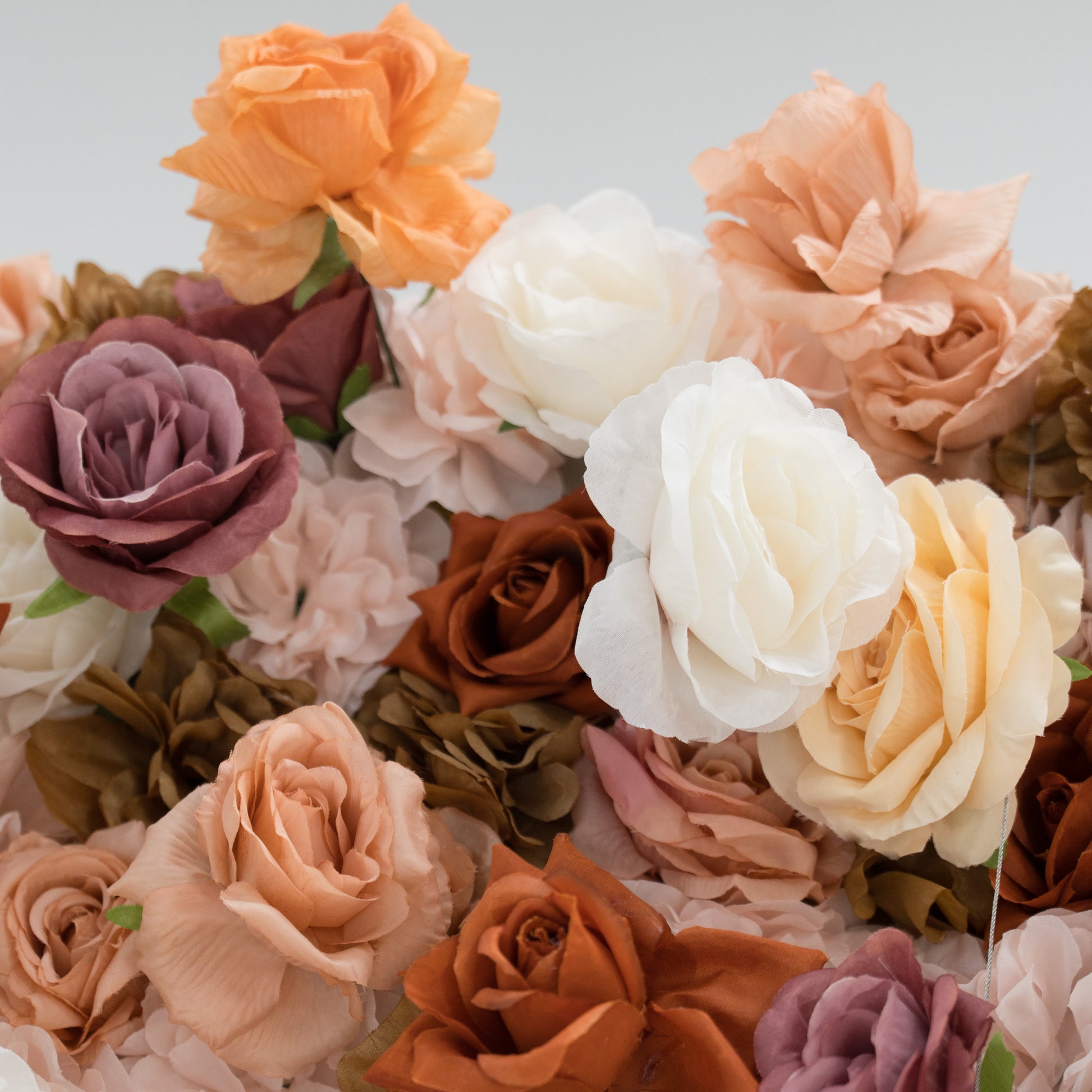 A close - up of artificial roses in soft, warm tones—peach, white, beige, brown, and purple. Their delicate, layered petals create a lush, textured arrangement, ideal for decorative backdrops or floral displays, evoking a romantic and elegant ambiance. 