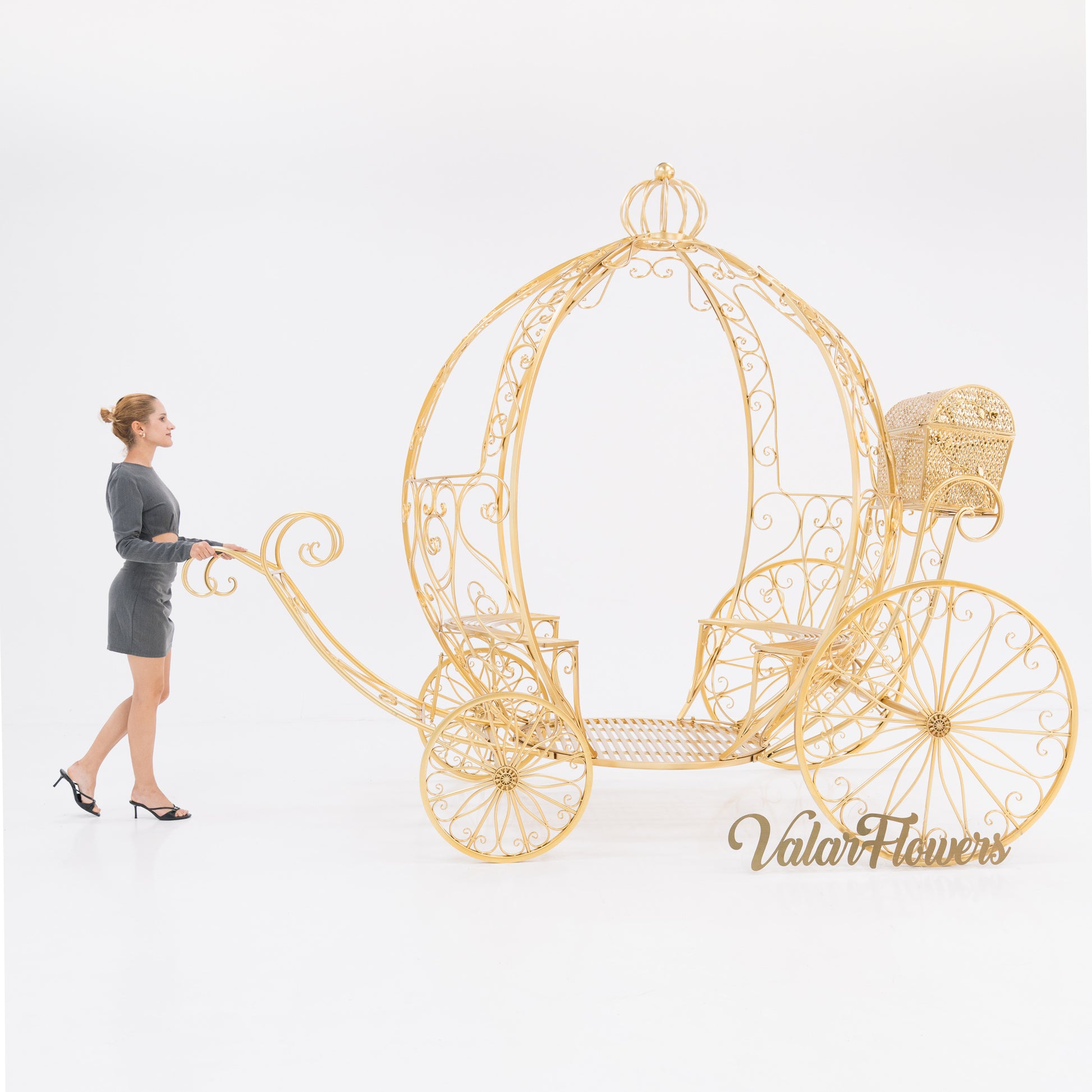 A woman in a gray dress and heels stands beside a large, gold - toned, ornate Cinderella - style carriage. Intricate wrought - iron scrollwork adorns the carriage, with a small latticework trunk. “ValarFlowers” branding adds a touch, perfect for fairy - tale - themed events. 