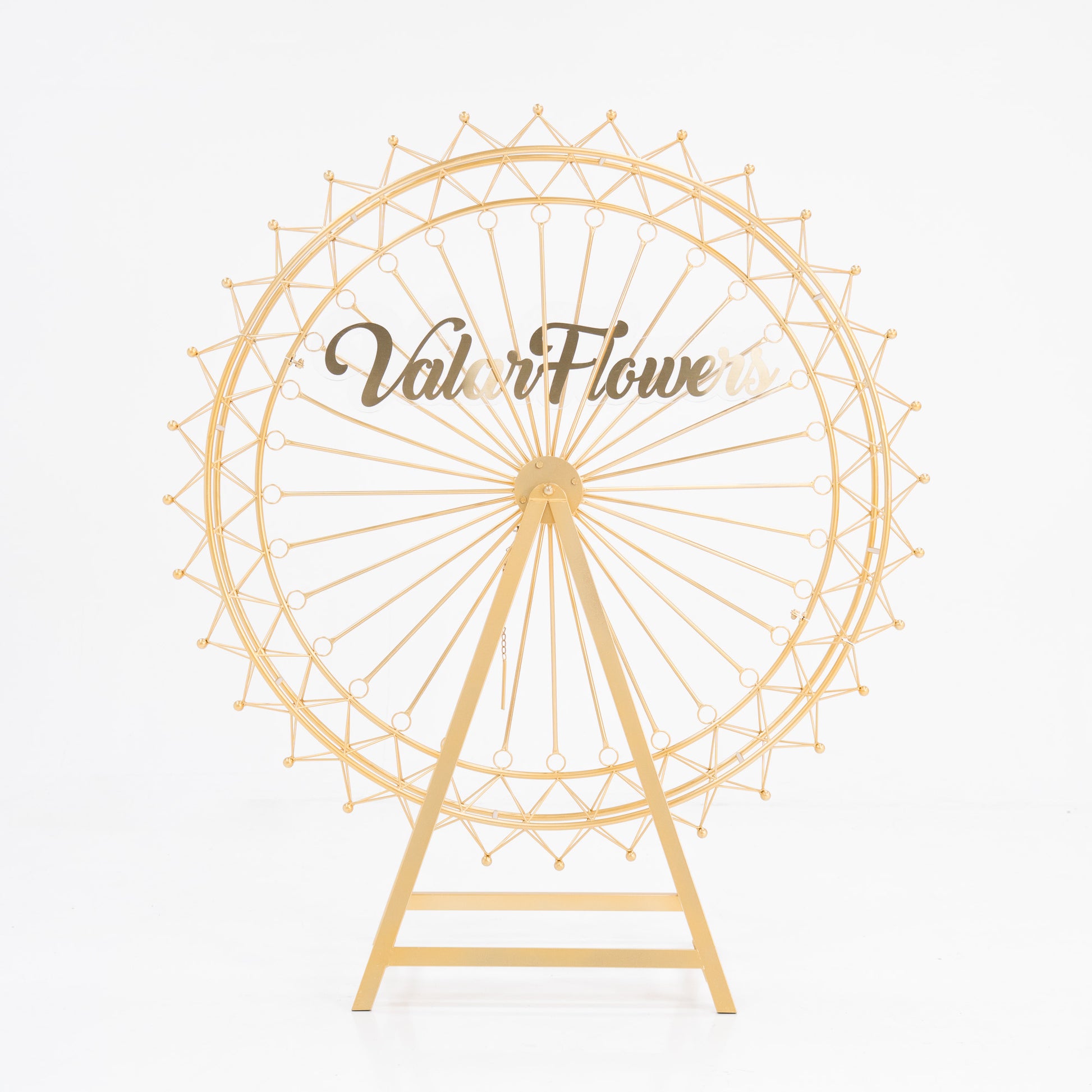 A golden, metal Ferris wheel decoration with a triangular stand. The wheel features intricate spokes and small spheres at the edges. In the center, a clear acrylic sign displays “ValarFlowers” in stylish, brown script. Ideal for decorative, branded event setups. 
