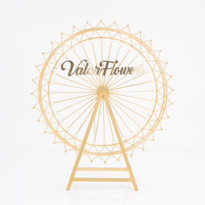 A golden, metal Ferris wheel decoration with a triangular stand. The wheel features intricate spokes and small spheres at the edges. In the center, a clear acrylic sign displays “ValarFlowers” in stylish, brown script. Ideal for decorative, branded event setups. 