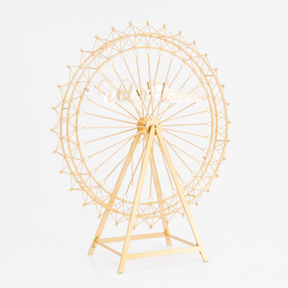 A golden, metal Ferris wheel decoration with a triangular stand. It features intricate spokes and small spheres along the rim. In the center, a stylish, cursive “ValarFlowers” sign adds branding. Ideal for decorative, themed event setups. 
