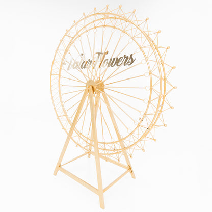A golden metal Ferris wheel decoration showcases an open, intricate lattice design. With a triangular stand and circular spokes, it forms a delicate, ornamental structure. The shimmering gold finish exudes elegance, ideal for decorative event setups, and has “ValarFlowers” branding on it. 
