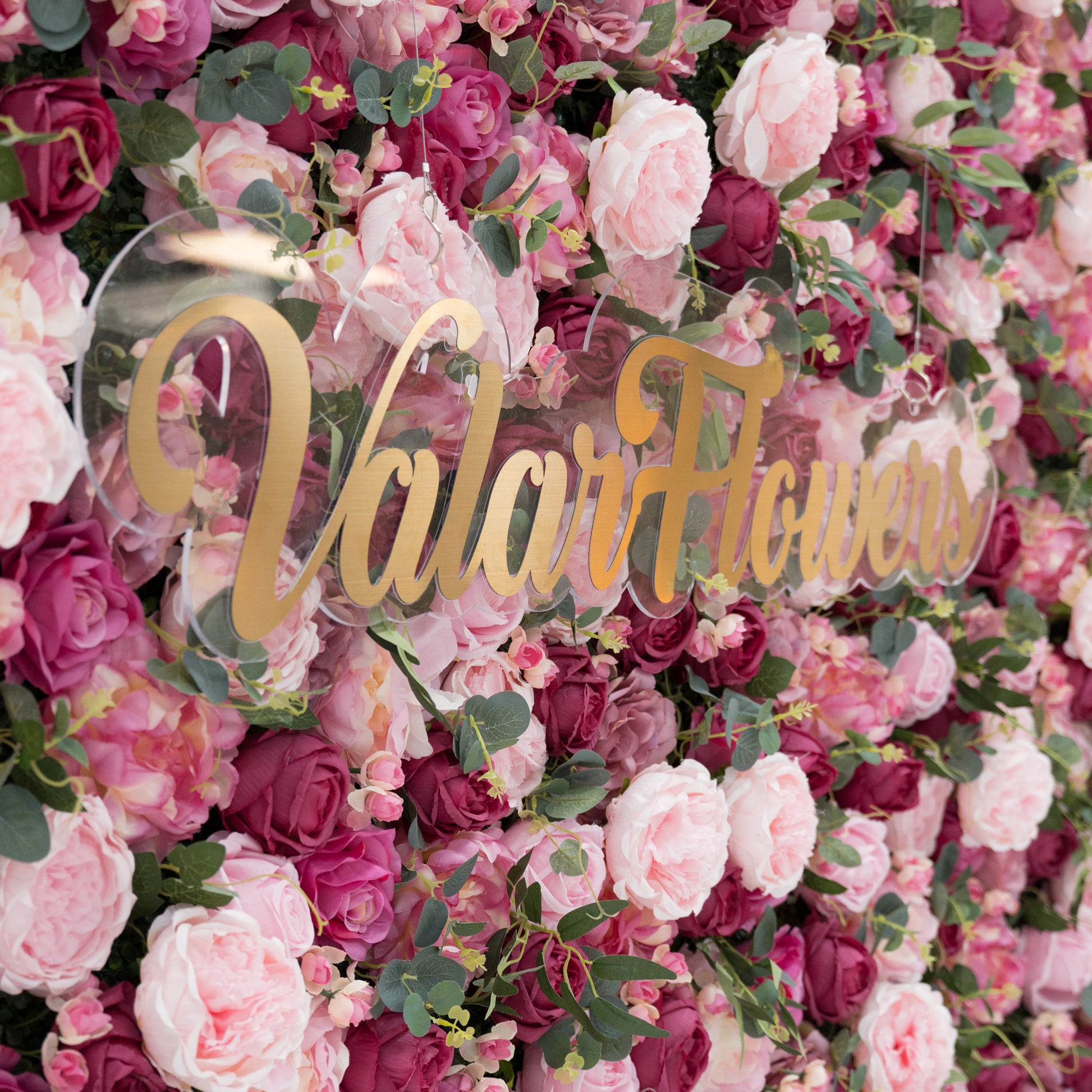 A lush artificial flower wall with pink and deep - pink blooms, green foliage. A clear acrylic sign with gold “ValarFlowers” lettering stands out, creating an elegant, branded backdrop—ideal for events, blending natural charm and sophistication. 