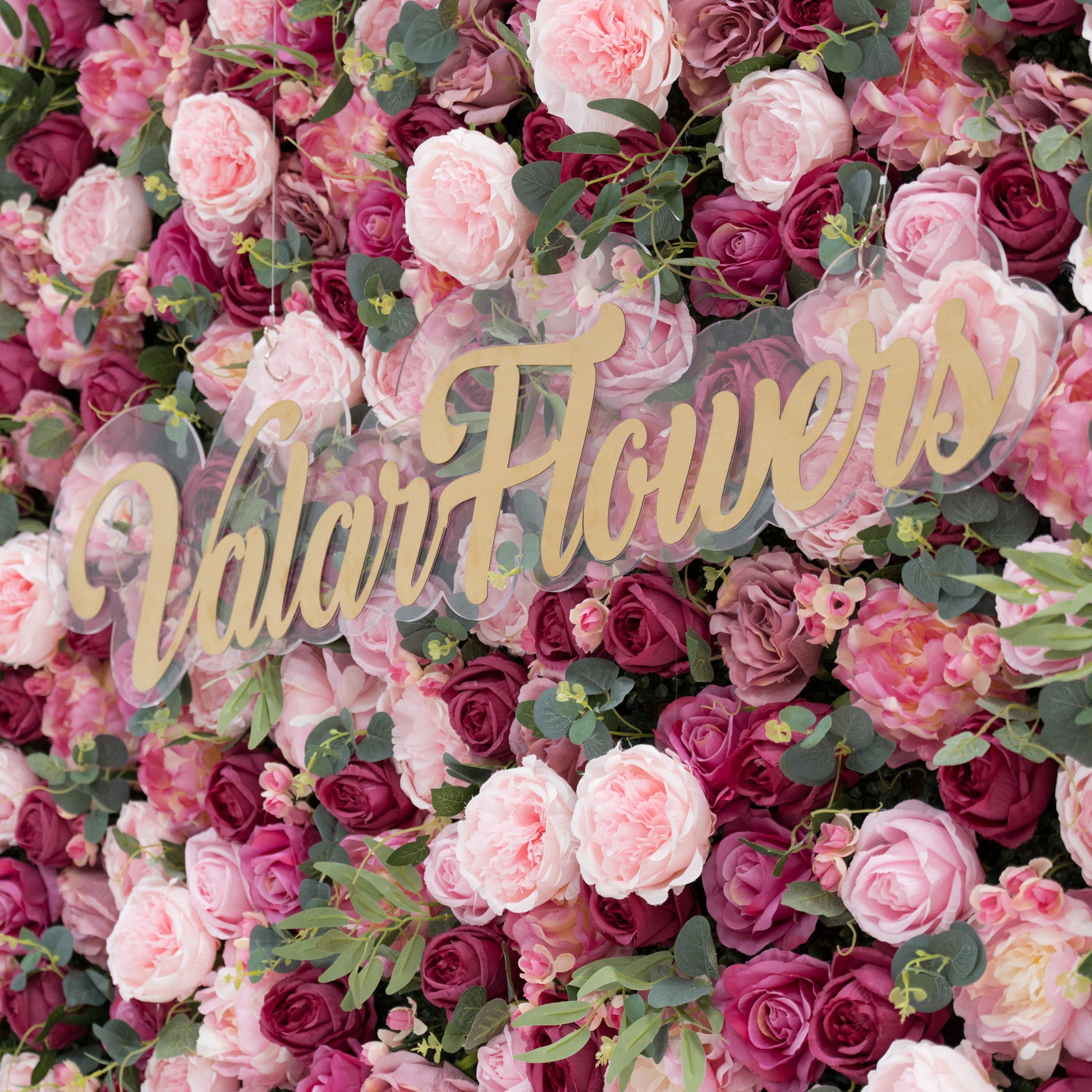 A lush artificial flower wall features pink and deep - pink roses, plus green foliage. A clear acrylic sign with gold “ValarFlowers” lettering stands out, creating an elegant, branded backdrop—ideal for events, blending natural charm and sophisticated branding. 