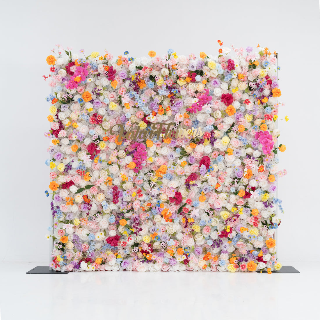 A large, square floral wall bursts with a vibrant mix of colorful artificial flowers—pink, yellow, orange, purple, and more. Dense clusters create a lush, lively display. With the “Valor Flowers” sign in the center, set against a plain white background and floor, it exudes a cheerful, festive vibe, perfect for events like weddings, parties, or as a decorative photo backdrop.