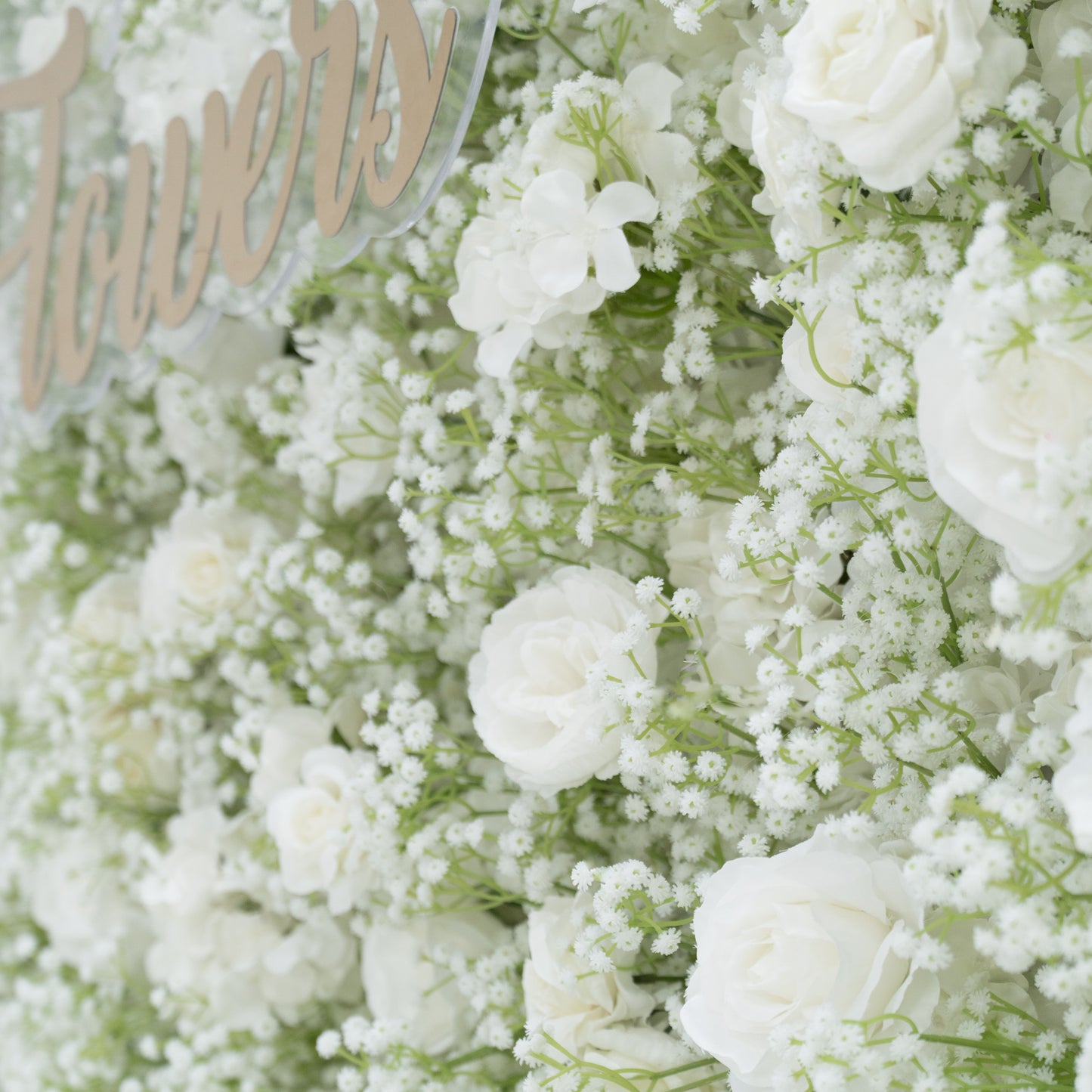 This image showcases a lush arrangement of white flowers, including roses and baby's breath, densely clustered together. Partially visible is a decorative sign with elegant lettering, adding a touch of sophistication. The blooms create a soft, pure, and elegant visual effect, exuding a sense of tranquility and beauty, perfect for decorative purposes in events or as a symbol of innocence and grace.