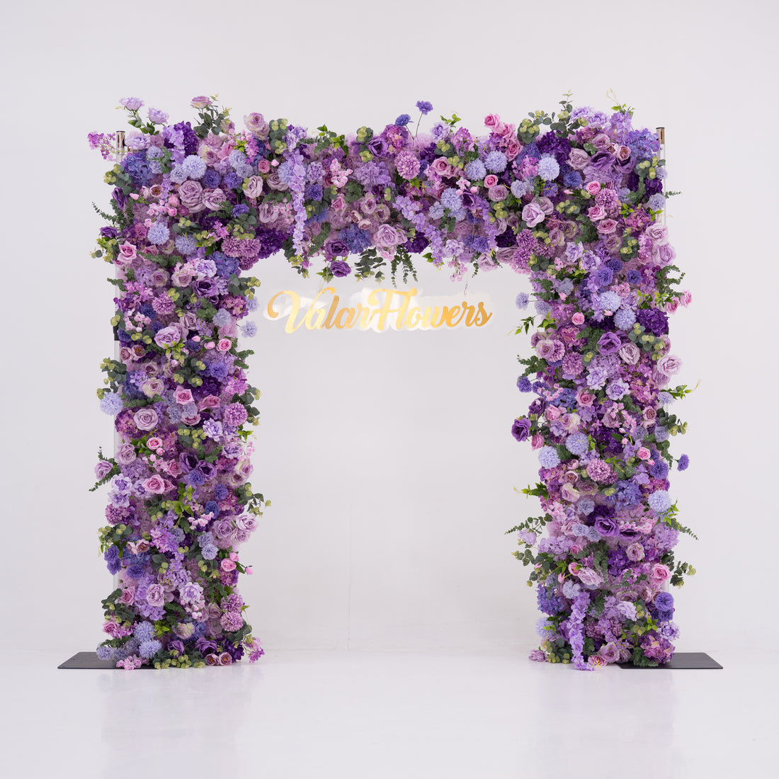 This image showcases a stunning floral arch by Valar Flowers, set against a plain white indoor background. It features a dense arrangement of artificial flowers in various shades of purple, lavender, and pink, creating an elegant, nature-inspired display. The brand’s name is elegantly integrated in gold, making it perfect for adding a luxurious, sophisticated element to events or interiors, highlighting Valar Flowers’ craftsmanship in eye-catching decor.