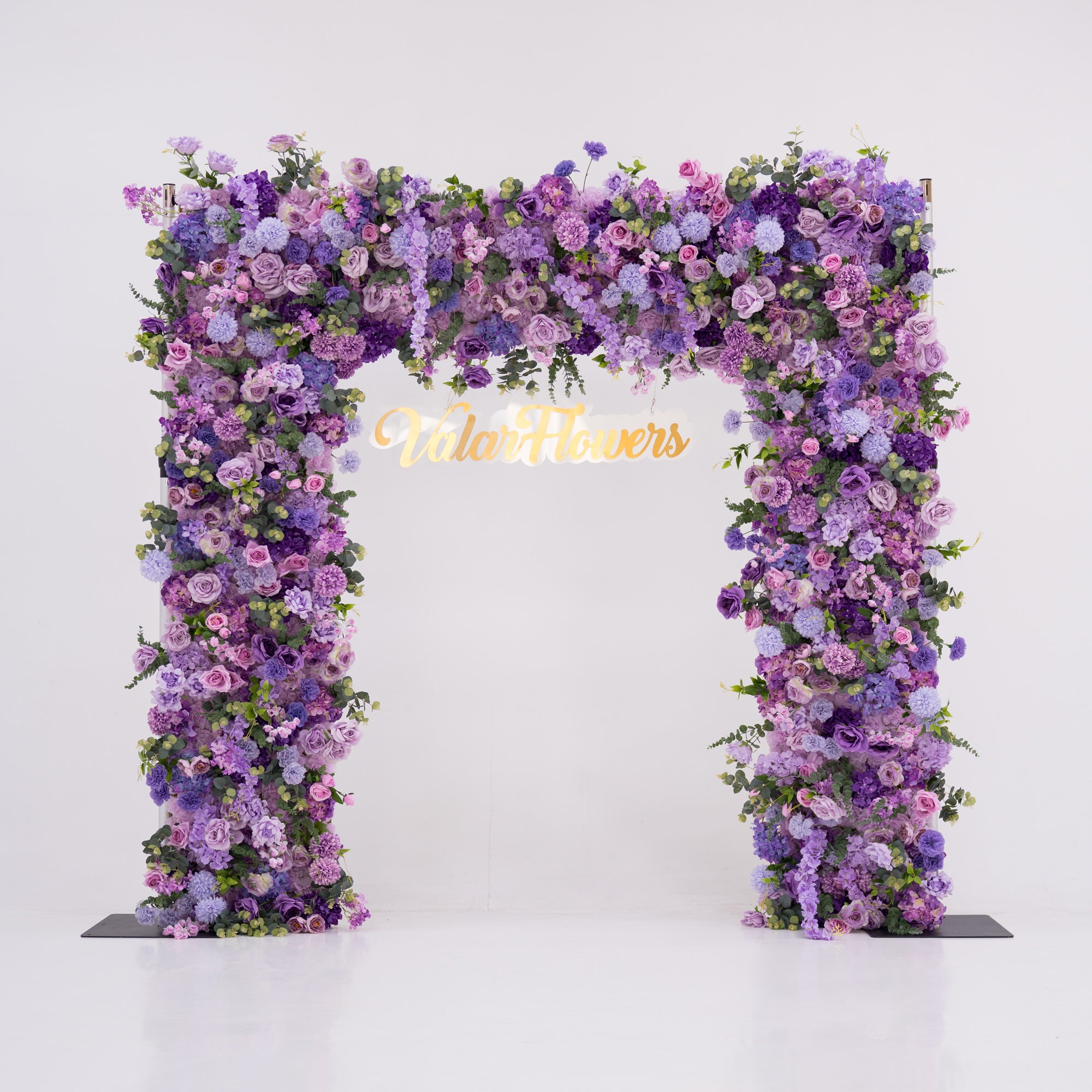 This image showcases a stunning floral arch by Valar Flowers, set against a plain white indoor background. It features a dense arrangement of artificial flowers in various shades of purple, lavender, and pink, creating an elegant, nature-inspired display. The brand’s name is elegantly integrated in gold, making it perfect for adding a luxurious, sophisticated element to events or interiors, highlighting Valar Flowers’ craftsmanship in eye-catching decor.