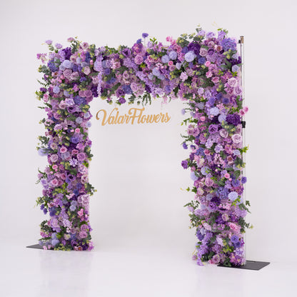 This image showcases a stunning floral arch by Valar Flowers, set against a plain white indoor background. It features a dense arrangement of artificial flowers in various shades of purple, lavender, and pink, creating an elegant, nature-inspired display. The brand’s name is elegantly integrated in gold, making it perfect for adding a luxurious, sophisticated element to events or interiors, highlighting Valar Flowers’ craftsmanship in eye-catching decor.