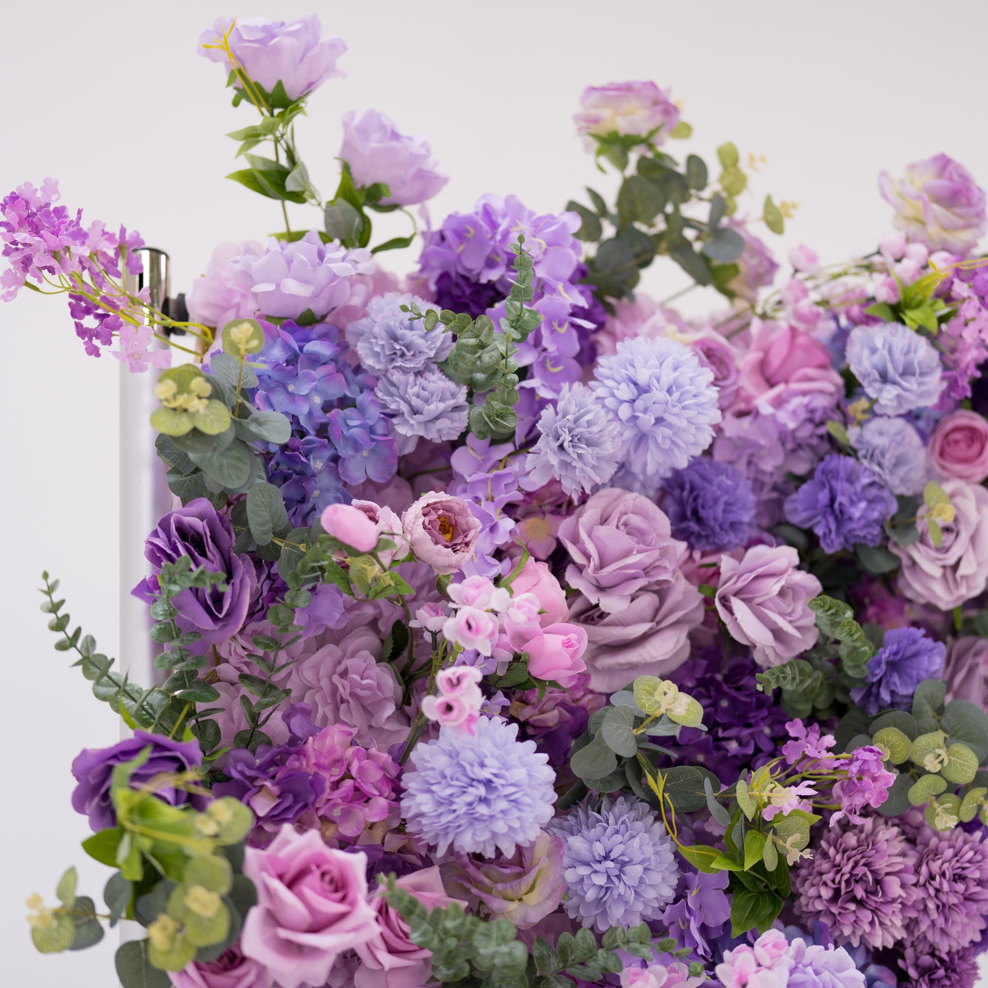 This image showcases a vibrant section of a floral backdrop by Valar Flowers, featuring a dense arrangement of artificial flowers in various shades of purple, lavender, and pink. The textured display includes varied blooms like roses, hydrangeas, and other floral varieties, complemented by green foliage. Exuding elegance and sophistication, it’s perfect for adding a luxurious, nature-inspired element to events or interiors, highlighting Valar Flowers’ craftsmanship in creating eye-catching, detailed decor.