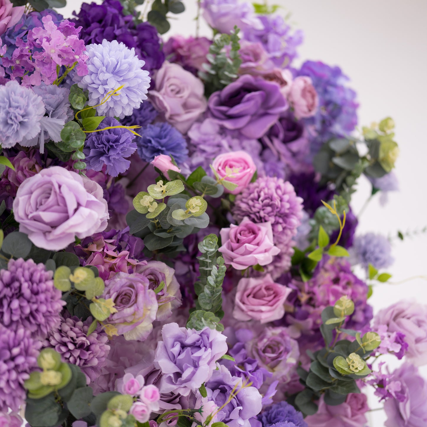 This image showcases a vibrant section of a floral backdrop by Valar Flowers, featuring a dense arrangement of artificial flowers in various shades of purple, lavender, and pink. The textured display includes roses, hydrangeas, and wisteria-like blooms, complemented by green foliage. 