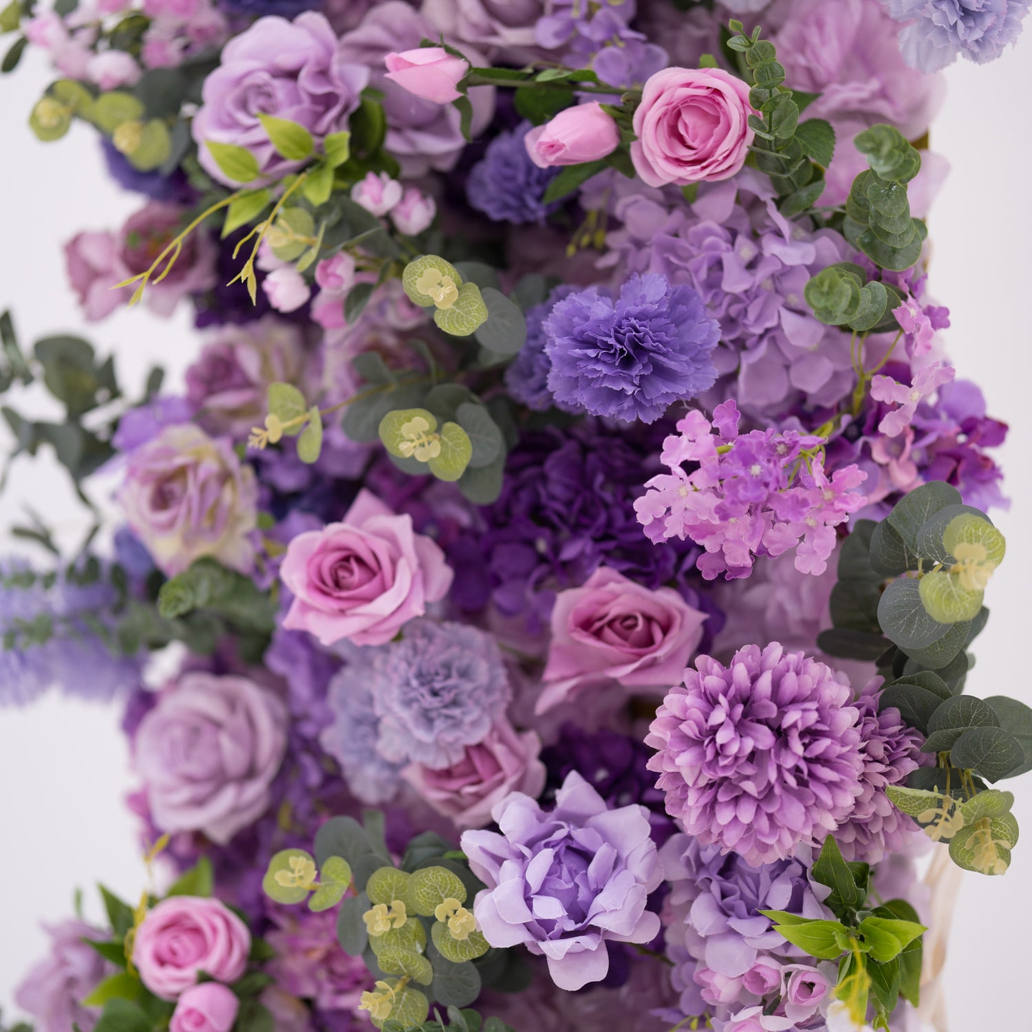 This image showcases a vibrant section of a floral backdrop by Valar Flowers, featuring a dense arrangement of artificial flowers in various shades of purple, lavender, and pink. The textured display includes roses, carnations, and other blooms, complemented by green foliage like eucalyptus. Exuding elegance, it’s perfect for adding a luxurious, nature-inspired element to events or interiors, highlighting Valar Flowers’ craftsmanship in detailed, eye-catching decor.