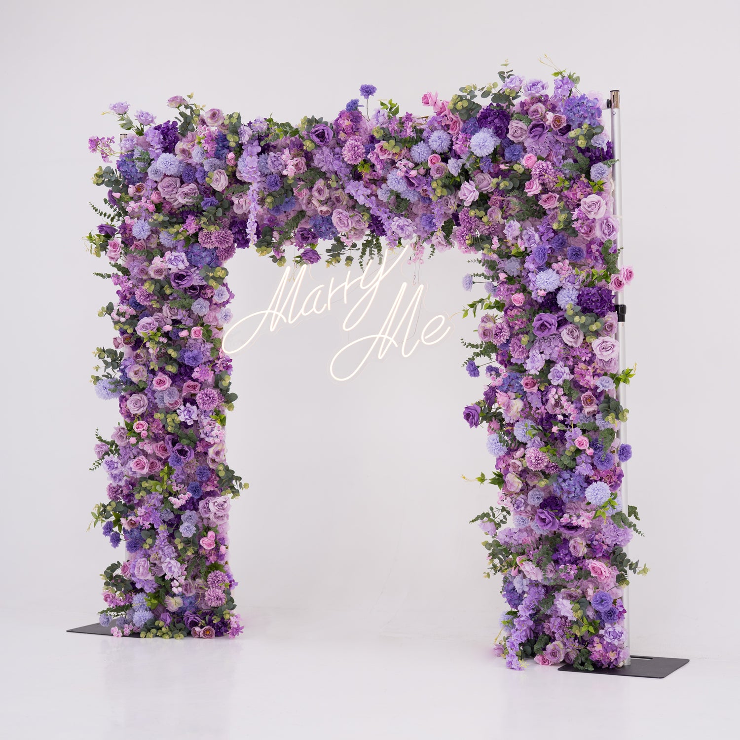 This image showcases a stunning floral arch by Valar Flowers, set against a plain white indoor background. It features a dense arrangement of artificial flowers in various shades of purple, lavender, and pink, with a “Marry Me” neon sign at the center. Exuding romance and elegance, it’s perfect for proposals or wedding events, highlighting Valar Flowers’ craftsmanship in creating luxurious, eye-catching decor.