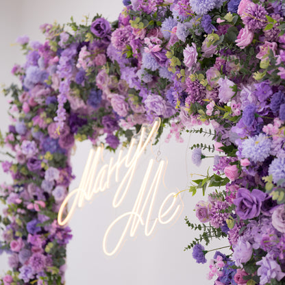 This image showcases a vibrant section of a floral arch by Valar Flowers, featuring a dense arrangement of artificial flowers in various shades of purple, lavender, and pink. A “Marry Me” neon sign is at the center, exuding romance. Perfect for proposals or weddings, it highlights Valar Flowers’ craftsmanship in creating luxurious, eye-catching decor.