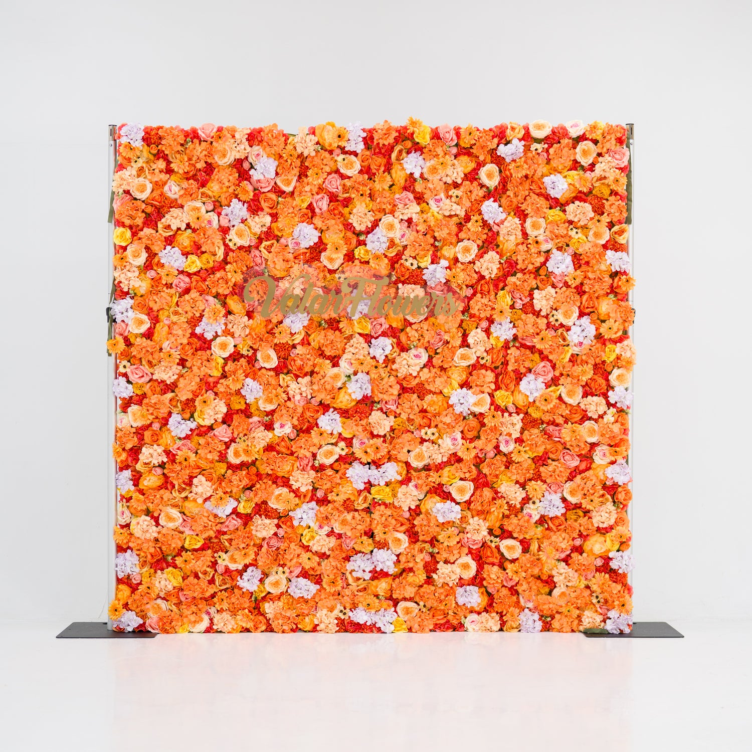 This image showcases a vibrant floral backdrop by Valar Flowers, featuring a dense arrangement of artificial flowers in warm shades of orange, peach, and hints of pink. The textured display exudes energy and warmth, with the brand’s name elegantly integrated. Set against a plain white indoor background, it’s perfect for adding a bold, nature-inspired element to events or interiors.