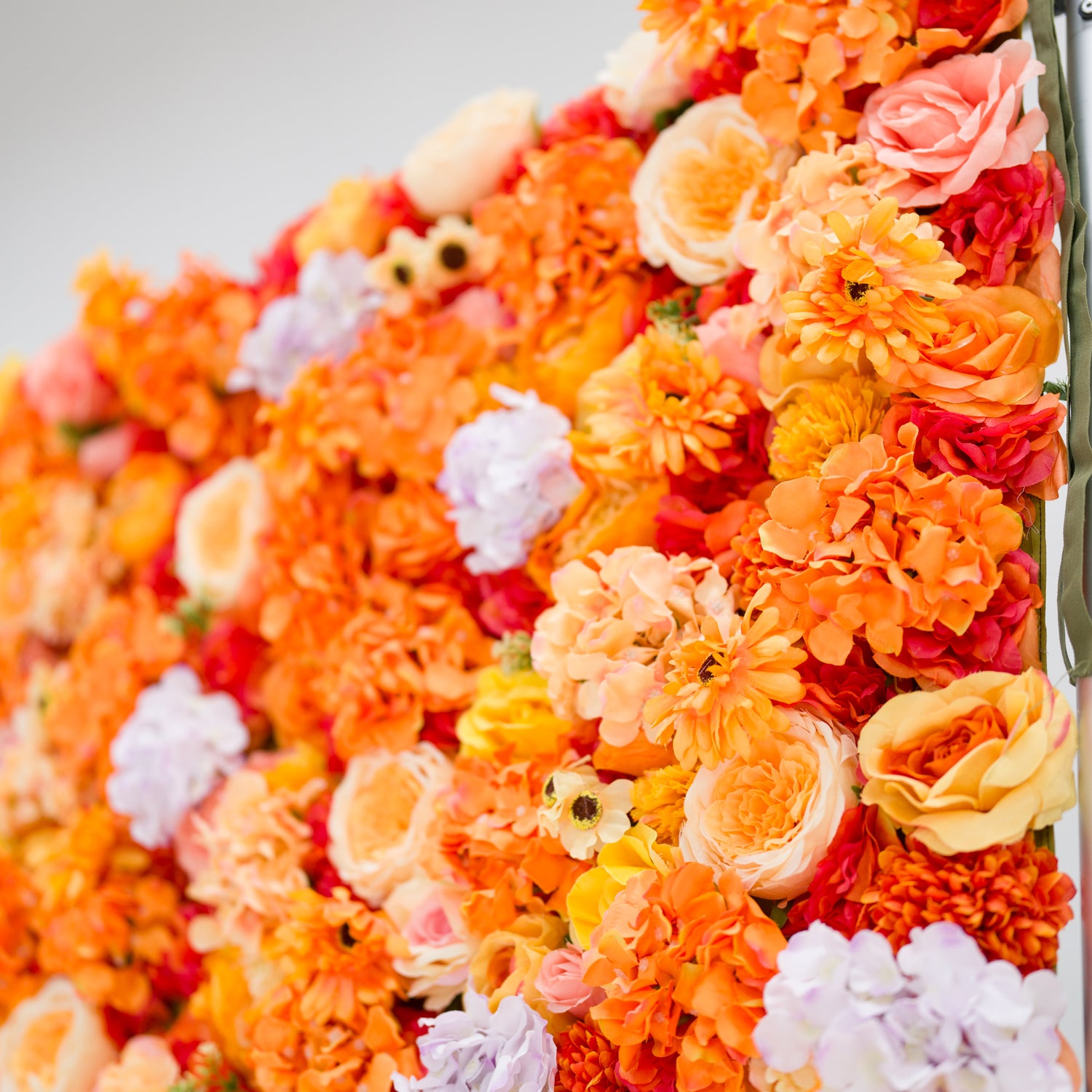 This image showcases a vibrant floral backdrop by Valar Flowers, featuring a dense arrangement of artificial flowers in warm shades of orange, peach, red, and hints of purple. The textured display, with varied blooms like roses and daisies, exudes energy and warmth. Perfect for adding a bold, nature-inspired element to events or interiors, it highlights the brand’s craftsmanship in creating eye-catching, sophisticated decor.