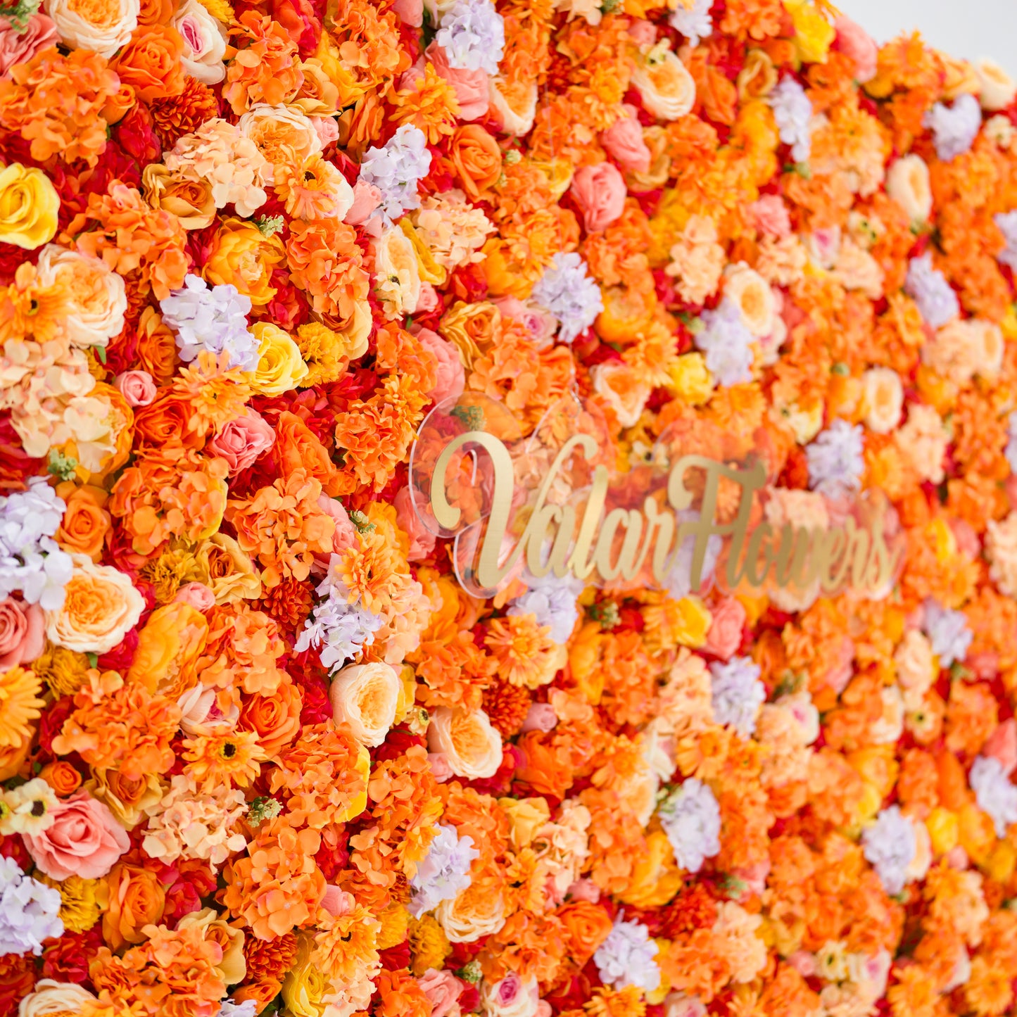 This image showcases a vibrant floral backdrop by Valar Flowers, featuring a dense arrangement of artificial flowers in warm shades of orange, peach, red, and hints of pink and purple. The textured display, with varied blooms like roses and daisies, exudes energy and warmth. The brand’s name is elegantly integrated in gold, making it perfect for adding a bold, nature-inspired element to events or interiors, highlighting Valar Flowers’ craftsmanship in eye-catching, sophisticated decor.