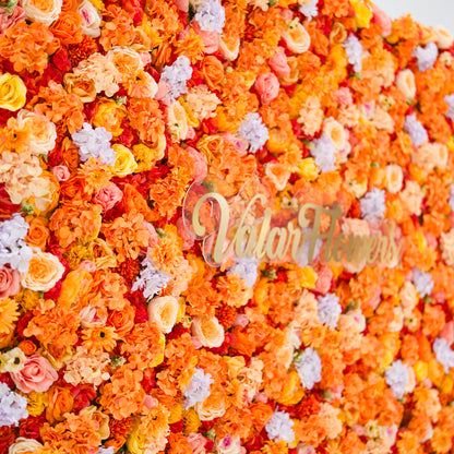This image showcases a vibrant floral backdrop by Valar Flowers, featuring a dense arrangement of artificial flowers in warm shades of orange, peach, red, and hints of pink and purple. The textured display, with varied blooms like roses and daisies, exudes energy and warmth. The brand’s name is elegantly integrated in gold, making it perfect for adding a bold, nature-inspired element to events or interiors, highlighting Valar Flowers’ craftsmanship in eye-catching, sophisticated decor.
