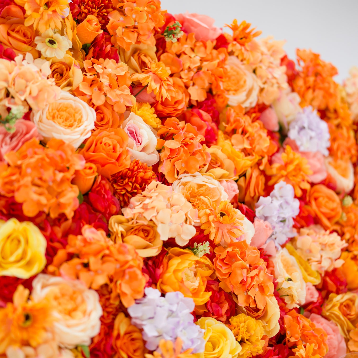 This image showcases a vibrant section of a floral backdrop by Valar Flowers, featuring a dense arrangement of artificial flowers in warm shades of orange, peach, red, yellow, and hints of purple. The textured display, with varied blooms like roses, hydrangeas, and daisies, exudes energy and warmth. Perfect for adding a bold, nature-inspired element to events or interiors, it highlights the brand’s craftsmanship in creating eye-catching, sophisticated decor.
