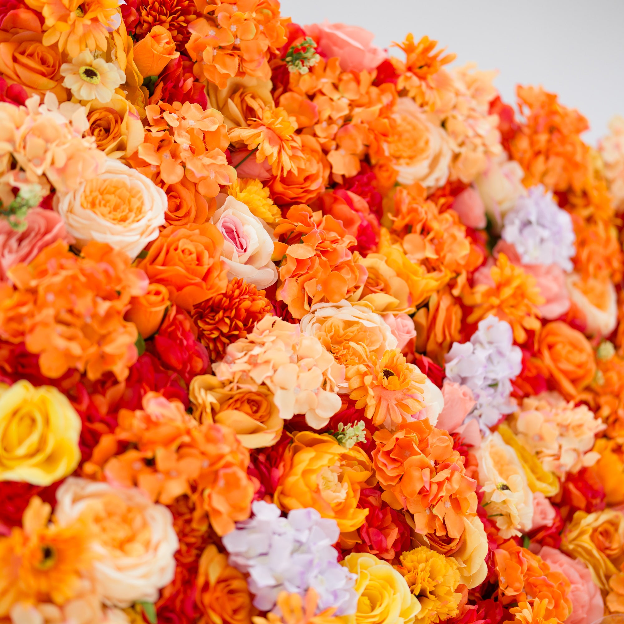 This image showcases a vibrant section of a floral backdrop by Valar Flowers, featuring a dense arrangement of artificial flowers in warm shades of orange, peach, red, yellow, and hints of purple. The textured display, with varied blooms like roses, hydrangeas, and daisies, exudes energy and warmth. Perfect for adding a bold, nature-inspired element to events or interiors, it highlights the brand’s craftsmanship in creating eye-catching, sophisticated decor.