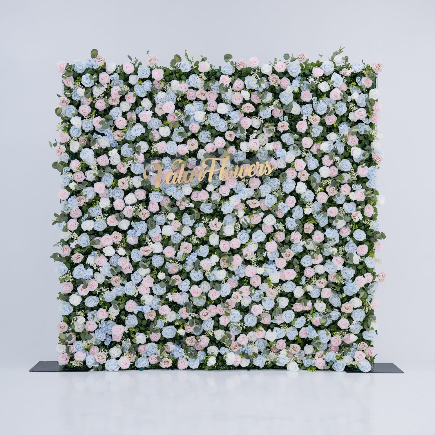 This image showcases a 2.4x2.4m (8x8ft) floral backdrop by Valar Flowers, set against a plain white indoor background. It features a dense arrangement of artificial flowers in various shades of blue, light blue, and pink, with the brand’s name elegantly integrated in gold. Exuding elegance, it’s perfect for adding a sophisticated, nature-inspired element to events, highlighting Valar Flowers’ craftsmanship in detailed, eye-catching decor.