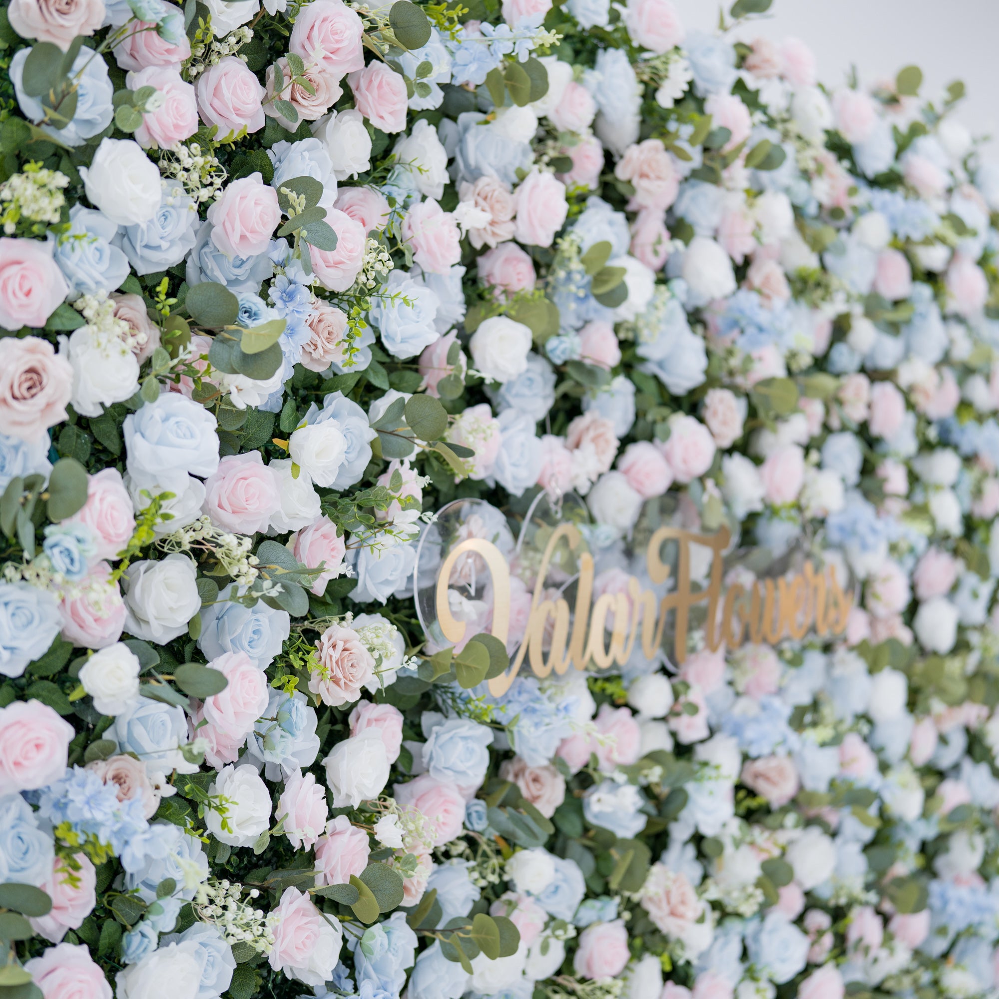 This image showcases a dense, vibrant backdrop of **artificial flowers** by Valar Flowers, featuring a harmonious mix of roses and blooms in shades of light blue, pink, white, and blush. Complemented by realistic green foliage, the textured arrangement exudes elegance. A gold “Valar Flowers” sign is integrated at the center, highlighting the brand’s craftsmanship. 
