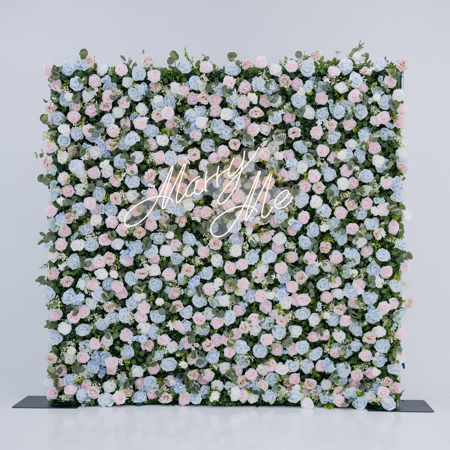 This image showcases a stunning floral backdrop featuring a dense arrangement of artificial flowers in soft shades of light blue, pink, and white, complemented by green foliage. At its center, a “Marry Me” neon sign adds a romantic touch. Set against a plain white indoor background, this elegant piece, ideal for proposals or weddings, highlights meticulous craftsmanship in creating sophisticated, eye-catching event decor.
