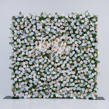 This image showcases a stunning floral backdrop featuring a dense arrangement of artificial flowers in soft shades of light blue, pink, and white, complemented by green foliage. At its center, a “Marry Me” neon sign adds a romantic touch. Set against a plain white indoor background, this elegant piece, ideal for proposals or weddings, highlights meticulous craftsmanship in creating sophisticated, eye-catching event decor.