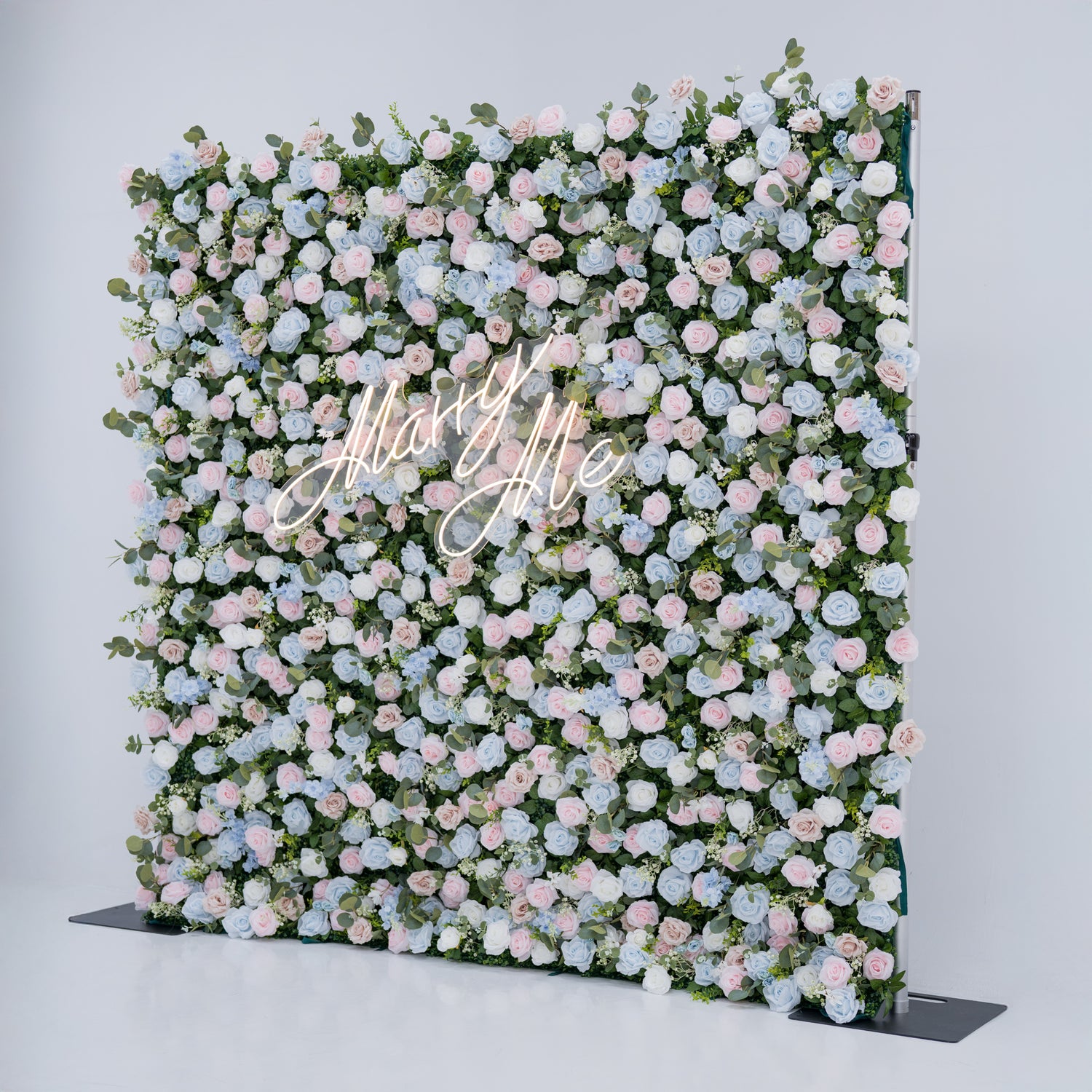 This image showcases a stunning floral backdrop with a dense arrangement of artificial flowers in soft shades of light blue, pink, and white, complemented by green foliage. At its center, a “Marry Me” neon sign adds a romantic touch. Set against a plain white indoor background, this elegant piece, ideal for proposals or weddings, highlights meticulous craftsmanship in creating sophisticated, eye-catching event decor.