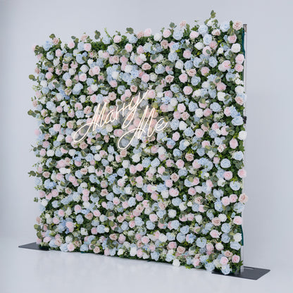 This image showcases a stunning floral backdrop with a dense arrangement of artificial flowers in soft shades of light blue, pink, and white, complemented by green foliage. At its center, a “Marry Me” neon sign adds a romantic touch. Set against a plain white indoor background, this elegant piece, ideal for proposals or weddings, highlights meticulous craftsmanship in creating sophisticated, eye-catching event decor.