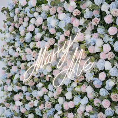 This image features a lush artificial floral backdrop with **roses** as the dominant flower type, in shades of light blue, pink, blush, and white. Accents include small blooms and green foliage like eucalyptus. At the center, a “Marry Me” neon sign adds romance. Ideal for proposals or weddings, it showcases meticulous craftsmanship in replicating realistic florals, blending various rose hues and textures for a sophisticated, eye-catching event decor piece.