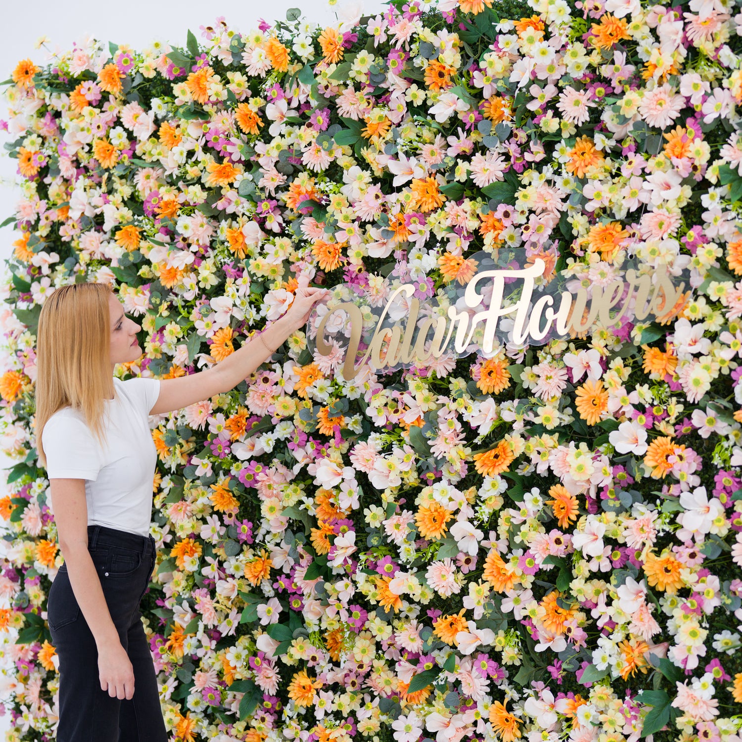 This close-up features a lush floral backdrop (bursting with pale pink, white, and orange daisy-like blooms, plus subtle purple accents) set against soft greenery. A woman (white top, black jeans) adjusts a polished "ValarFlowers" sign, adding a dynamic styling touch. The bright, varied palette and dense, vibrant blooms evoke a cheerful, fresh vibe—ideal for outdoor events, wedding decor, or festive photo backdrops.