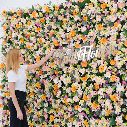 This close-up features a lush floral backdrop (bursting with pale pink, white, and orange daisy-like blooms, plus subtle purple accents) set against soft greenery. A woman (white top, black jeans) adjusts a polished "ValarFlowers" sign, adding a dynamic styling touch. The bright, varied palette and dense, vibrant blooms evoke a cheerful, fresh vibe—ideal for outdoor events, wedding decor, or festive photo backdrops.