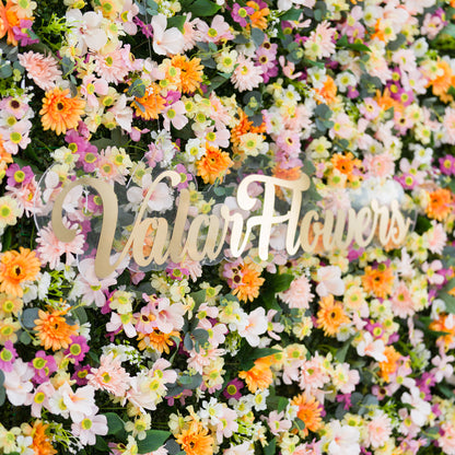 This intimate close-up showcases a lush floral arrangement: dense clusters of pale pink, white, and warm orange daisy-like blooms, accented with subtle purple flowers and soft greenery. A polished "ValarFlowers" sign (cursive, metallic) anchors the center, blending seamlessly with the vibrant, varied petals. The bright, layered palette evokes a cheerful, fresh vibe—ideal for event decor, wedding styling, or festive photo details.