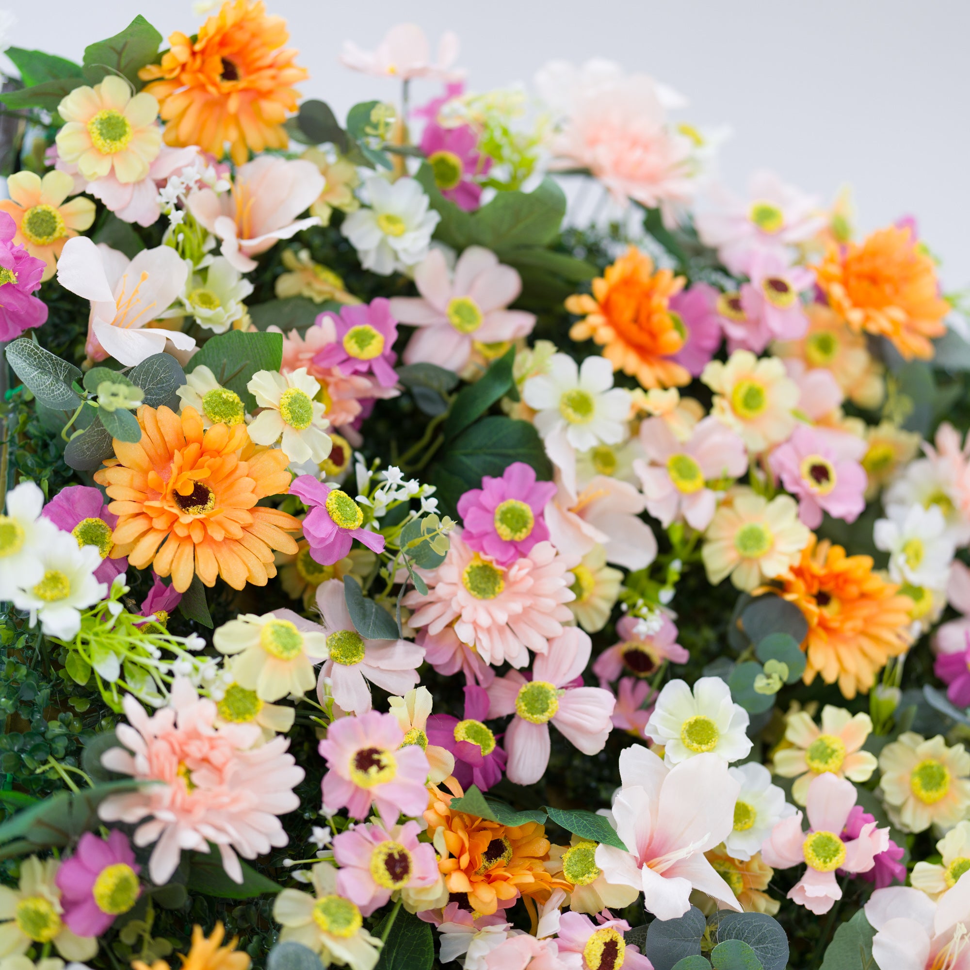 This detailed close-up bursts with a vibrant floral arrangement: lush clusters of warm orange daisies, soft pale pink blooms, crisp white flowers, and subtle purple accents, all centered on bright yellow-green cores. Lush green leaves and tiny filler blooms add texture, creating a dense, cheerful display. The varied pastel-and-orange palette evokes a fresh, lively vibe—ideal for event decor, wedding styling, or festive floral details.