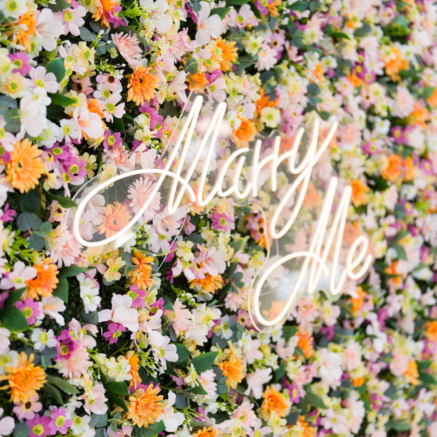This close-up features a lush floral backdrop: dense clusters of warm orange daisies, soft pale pink blooms, crisp white flowers, and subtle purple accents, all anchored by a glowing "Marry Me" neon sign (cursive, light-lit). Lush green leaves add texture, while the bright pastel-and-orange palette evokes a romantic, cheerful vibe—ideal for proposal setups, wedding decor, or intimate event styling.