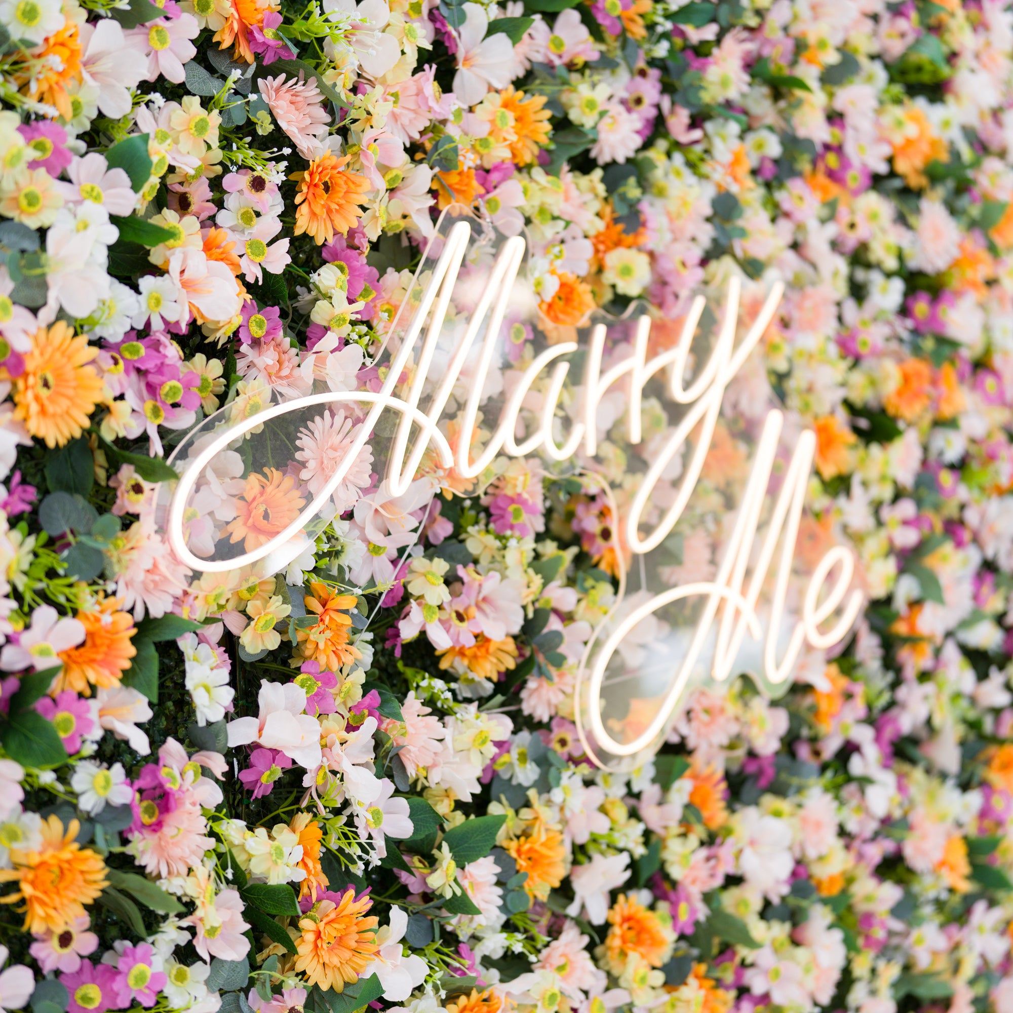This close-up features a lush floral backdrop: dense clusters of warm orange daisies, soft pale pink blooms, crisp white flowers, and subtle purple accents, all anchored by a glowing "Marry Me" neon sign (cursive, light-lit). Lush green leaves add texture, while the bright pastel-and-orange palette evokes a romantic, cheerful vibe—ideal for proposal setups, wedding decor, or intimate event styling.